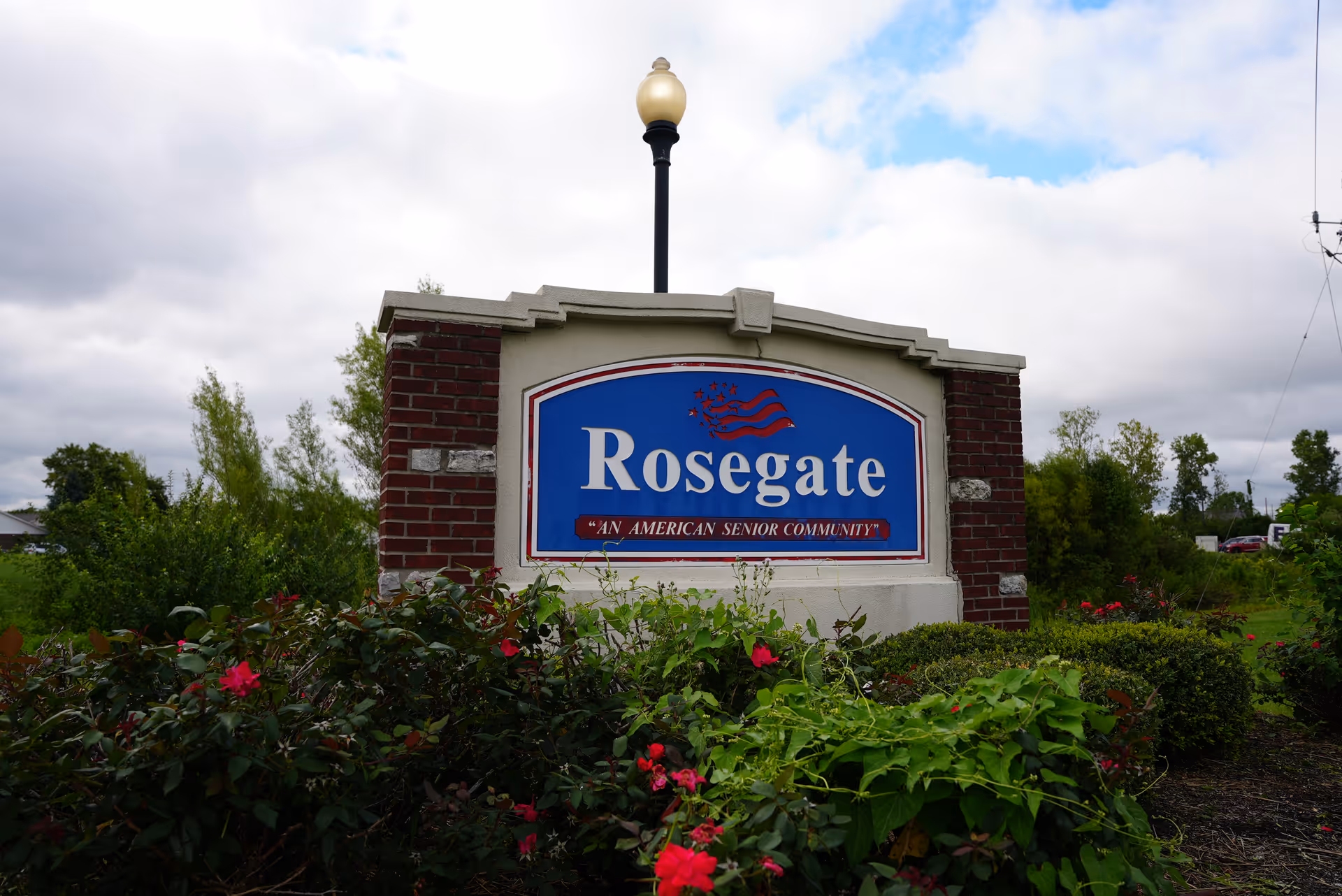 Brick and stone entrance sign reading "Rosegate" with landscaping and a lamppost under a cloudy sky.