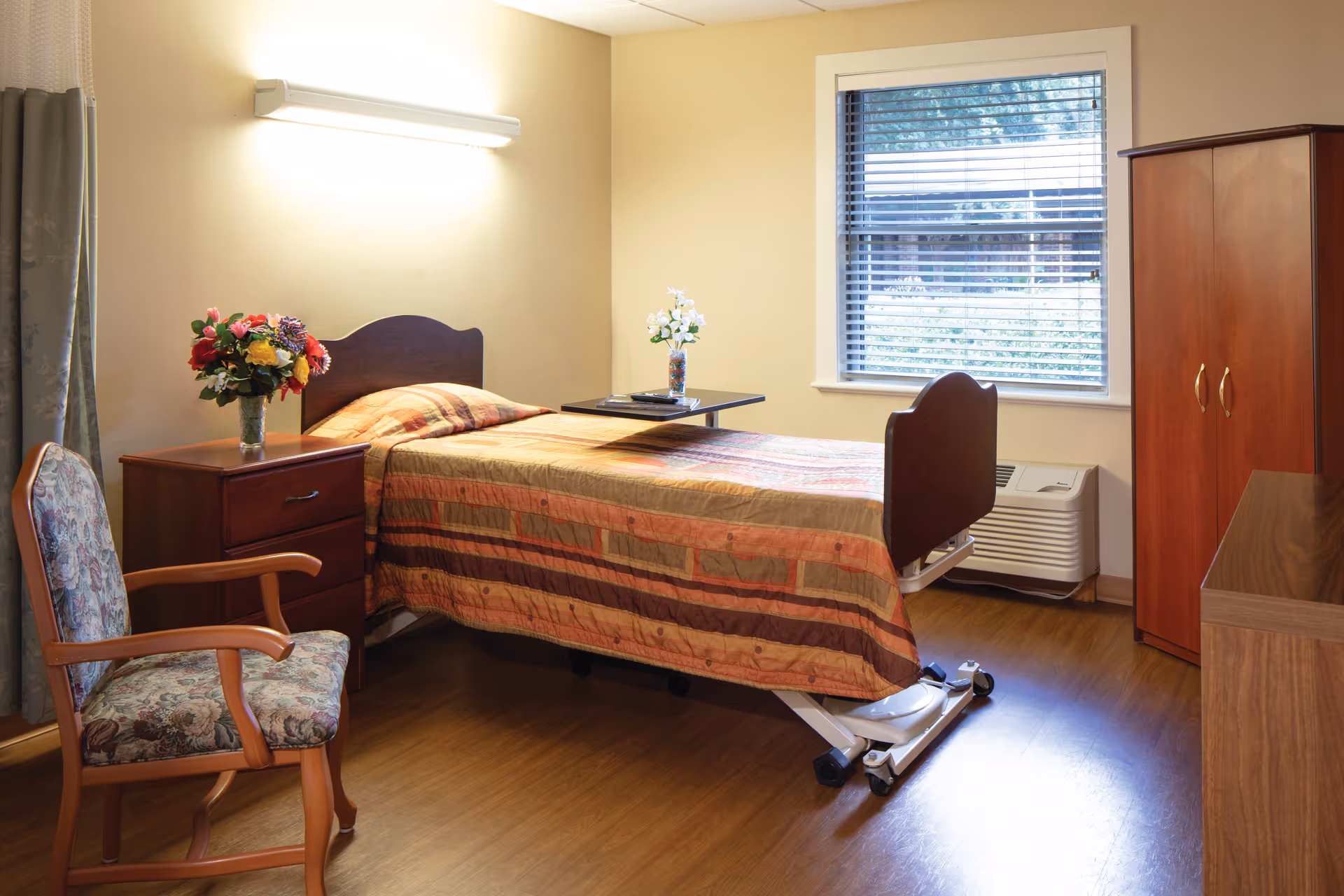 Bright patient bedroom with a bed, bedside table, chair, wardrobe, and window.
