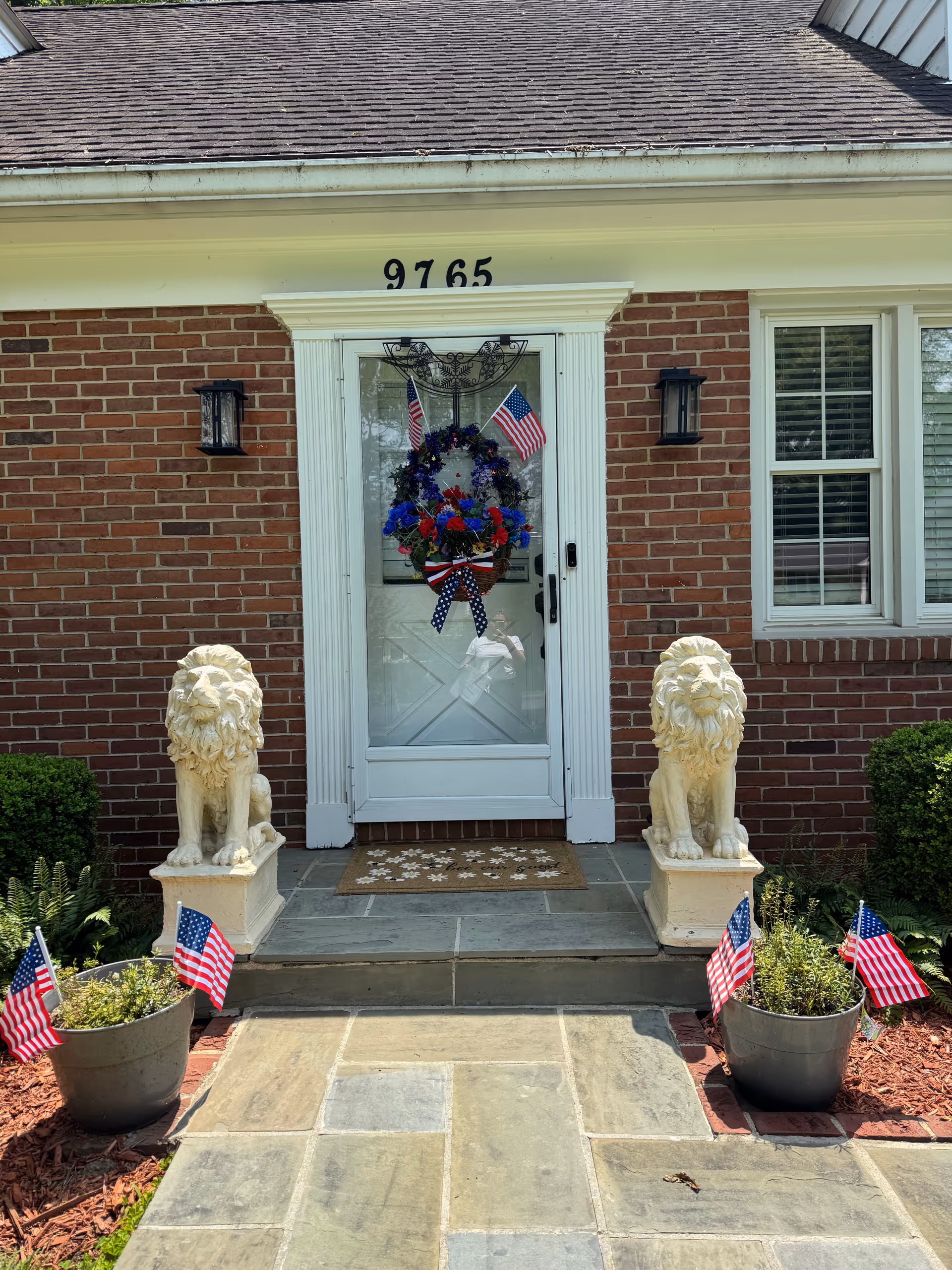 Brick assisted living entrance with wreath, American flags, lion statues, and potted plants.