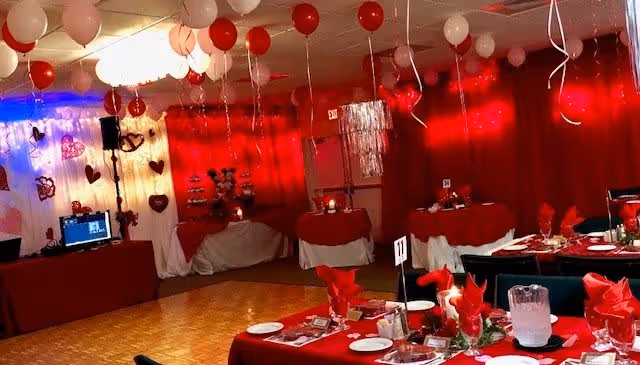 A decorated event room with red and white balloons hanging from the ceiling, red tablecloths, and tables set with plates, glasses, and red napkins. The walls are adorned with red curtains and heart-shaped decorations, creating a festive atmosphere.