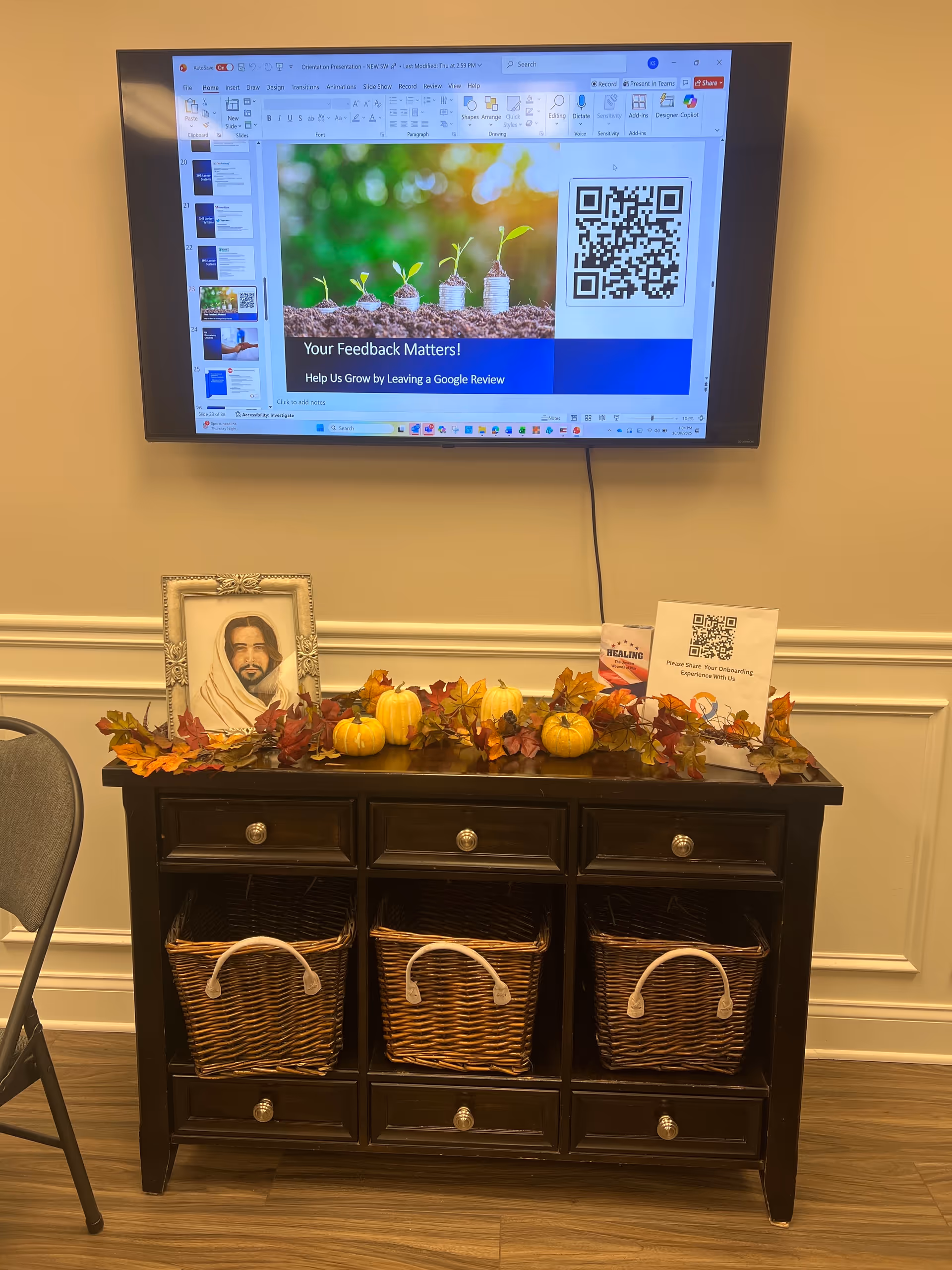 Indoor reception area with a dark wood cabinet, pumpkins, fall foliage, and a mounted presentation screen.
