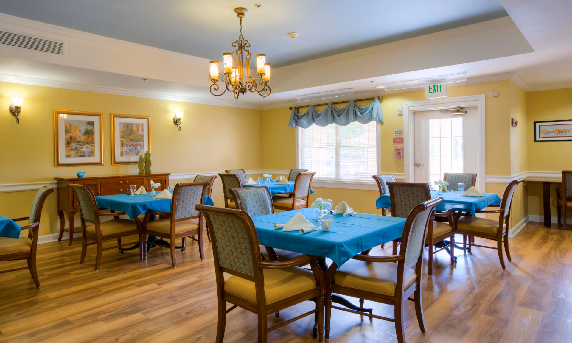 Dining room with turquoise-tablecloth tables and wooden chairs in a senior living facility.