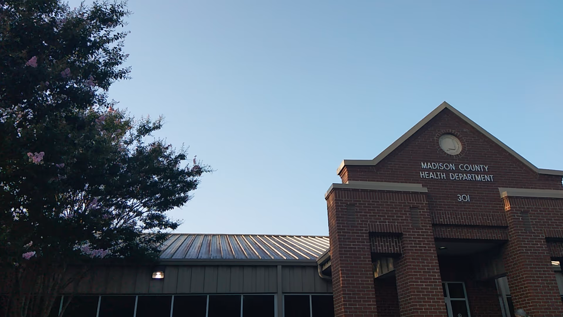 Brick Madison County Health Department building with a peaked front against a blue sky.