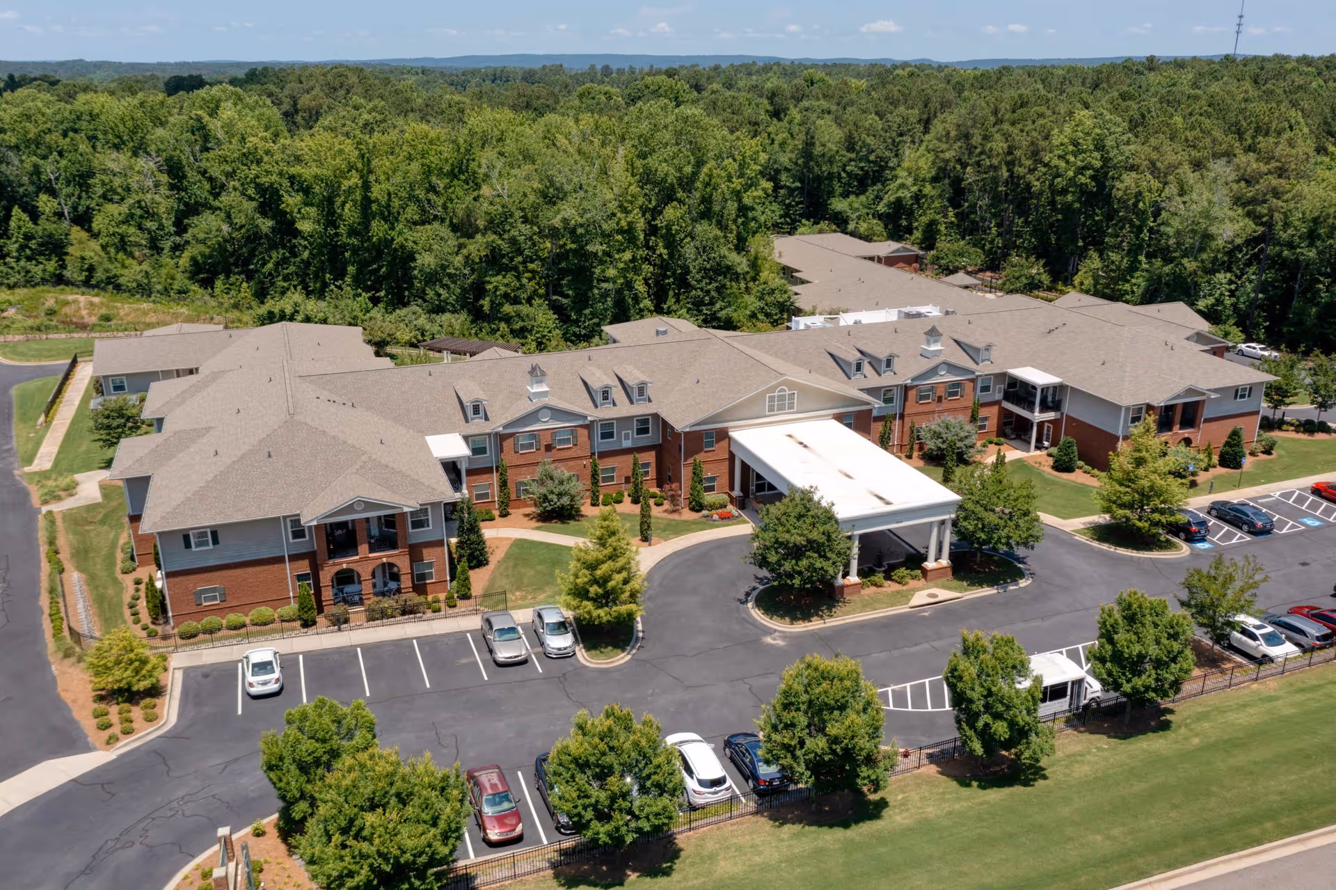 Aerial view of Camellia Walk of Evans senior living facility showing a large two-story building with a covered entrance, surrounded by parking lots, trees, and greenery with a forested area in the background under a partly cloudy sky.