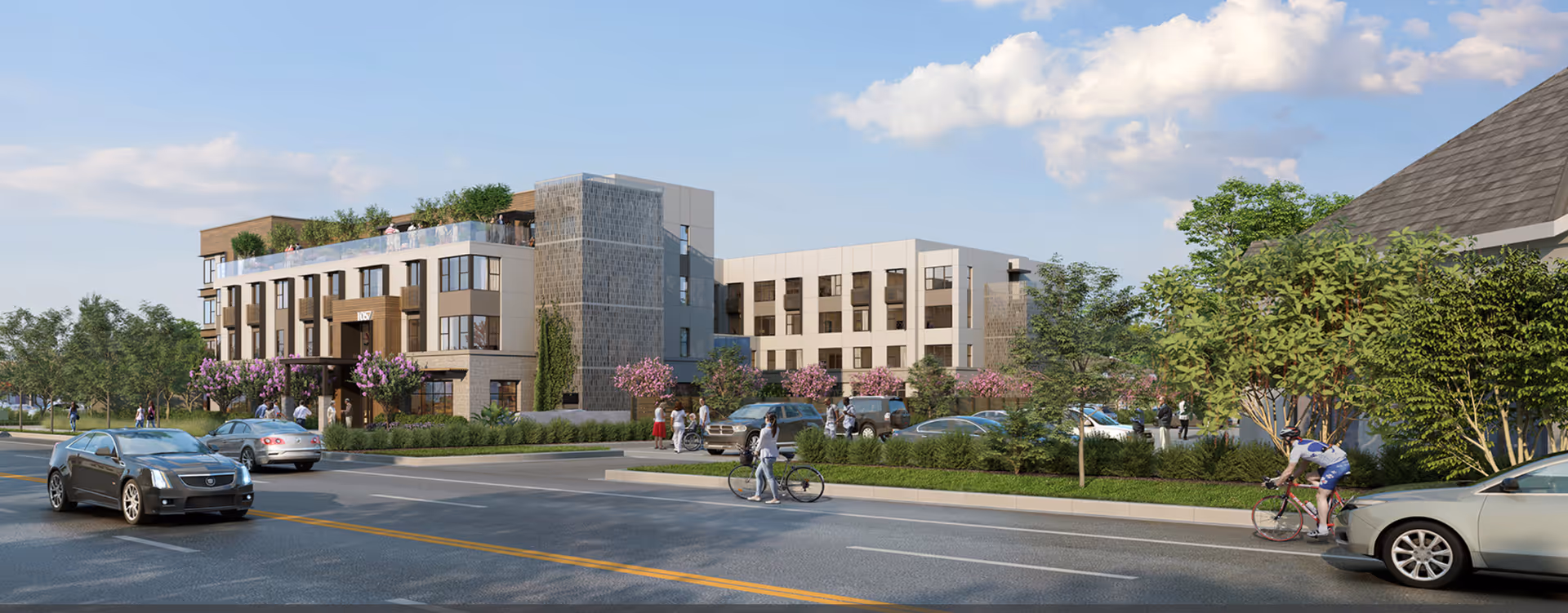 Exterior view of a modern multi-story assisted living and memory care facility with landscaped greenery, parked cars, people walking and biking, and a partly cloudy sky.