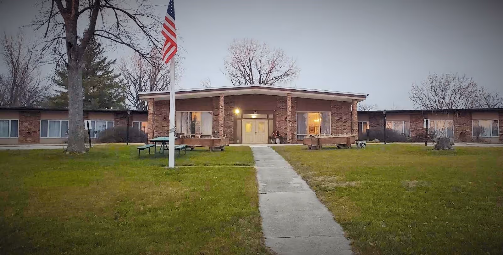 Front view of a single-story brick senior care building with a central entrance, flagpole, walkway, and lawn.