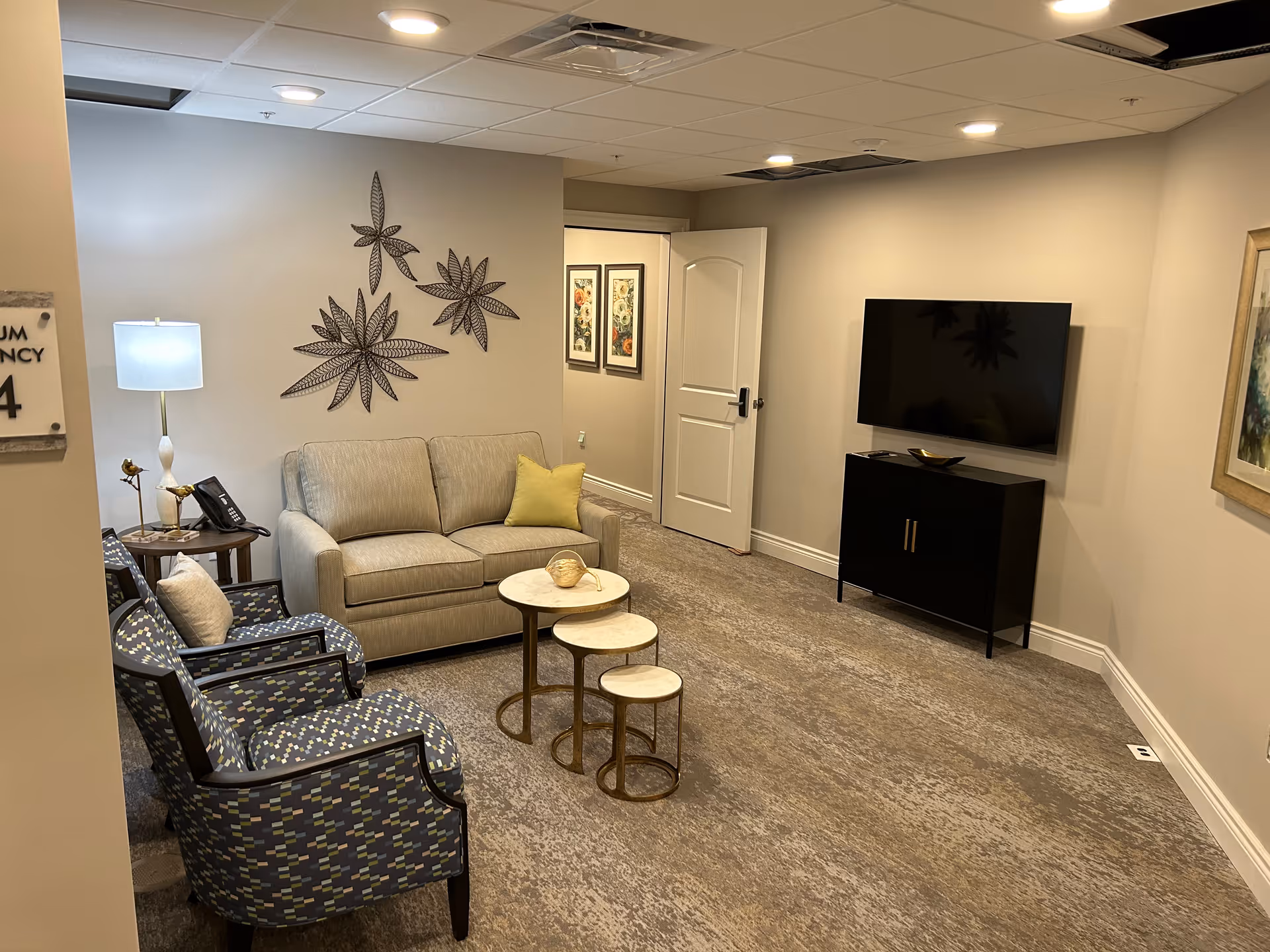 A cozy living room area with a beige sofa, two patterned armchairs, a set of three nesting round tables, a floor lamp, and a wall-mounted flat screen TV above a black cabinet. The walls are decorated with metal leaf art and framed pictures. The room has a carpeted floor and a white door slightly ajar leading to another room.
