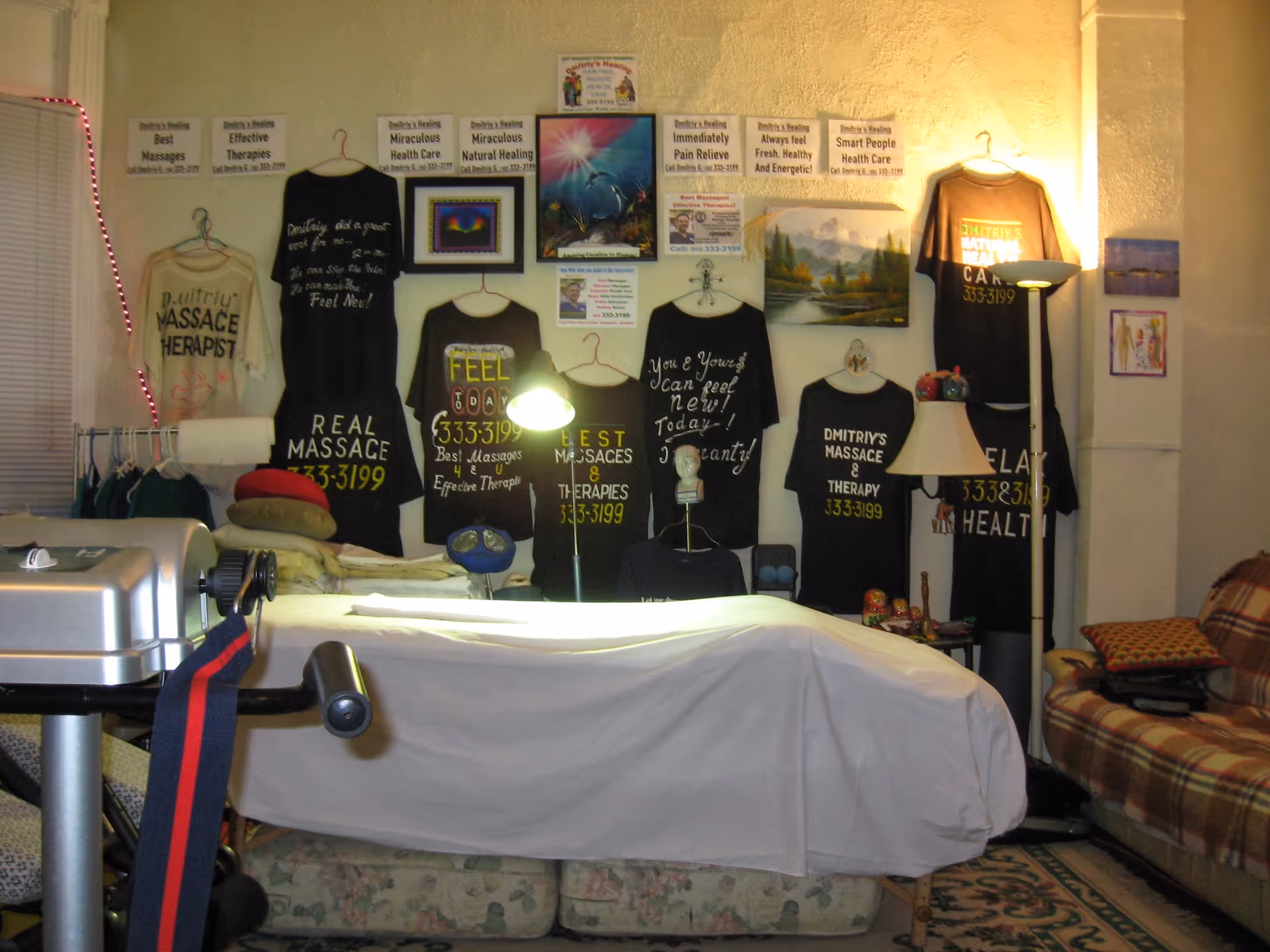 A cluttered interior with a massage table, wall-mounted shirts, posters, and a sofa.