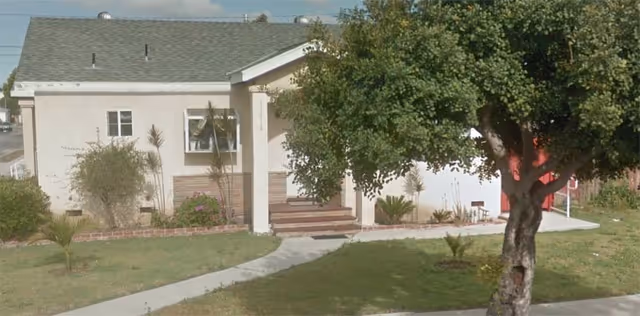 Front exterior of a single-story senior living facility with walkway, lawn, and trees.