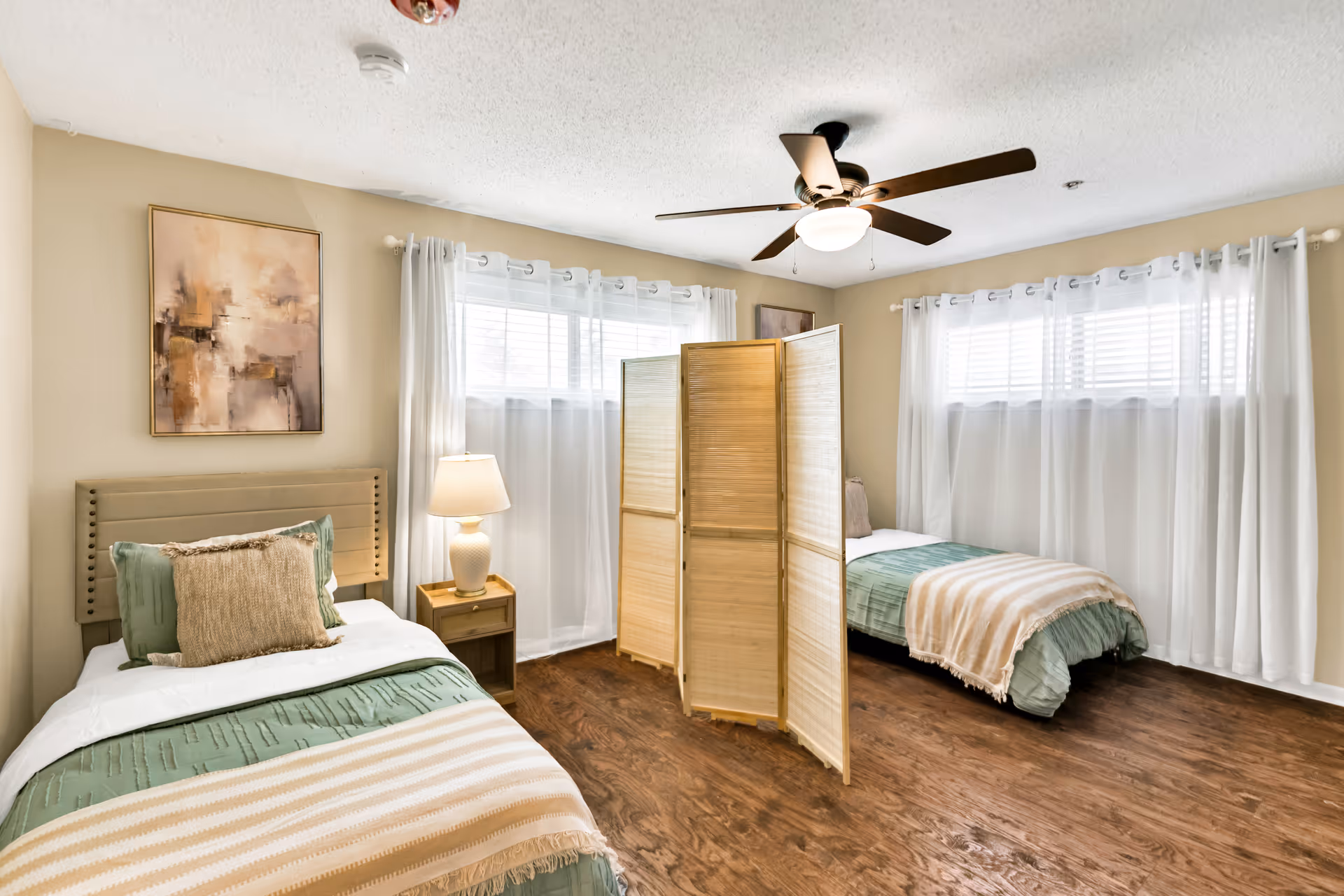 Two single beds with a folding screen, lamp, and windows with sheer curtains.