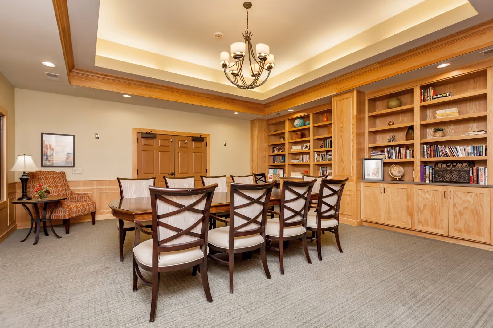 A well-lit room with a long wooden table surrounded by eight cushioned chairs with wooden frames. The room features built-in wooden bookshelves filled with books and decorative items along one wall. There is a patterned armchair with a small side table and lamp in one corner. The ceiling has recessed lighting and a chandelier with multiple lights. The walls are painted a light beige color with wooden trim and double wooden doors at the back.
