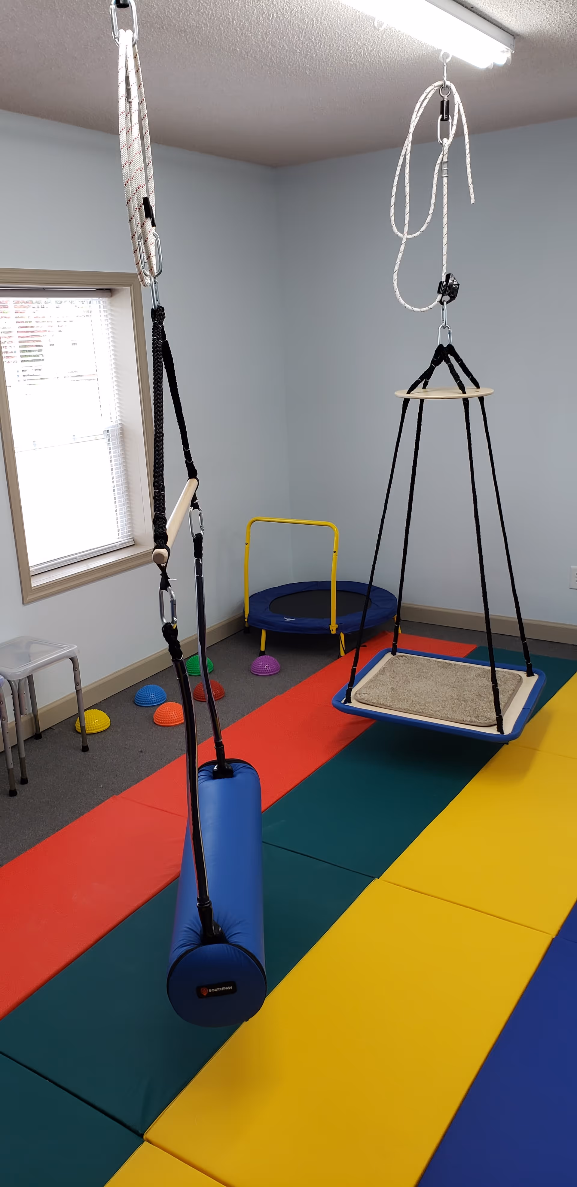 Indoor therapy room with hanging swings and colorful padded floor mats.