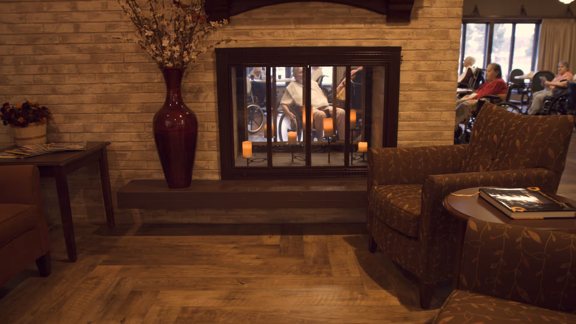 A cozy seating area in a senior living facility with a brown patterned armchair, a small round table with books, a fireplace with candles inside, and a large vase with decorative branches. In the background, elderly residents are seated, some in wheelchairs.