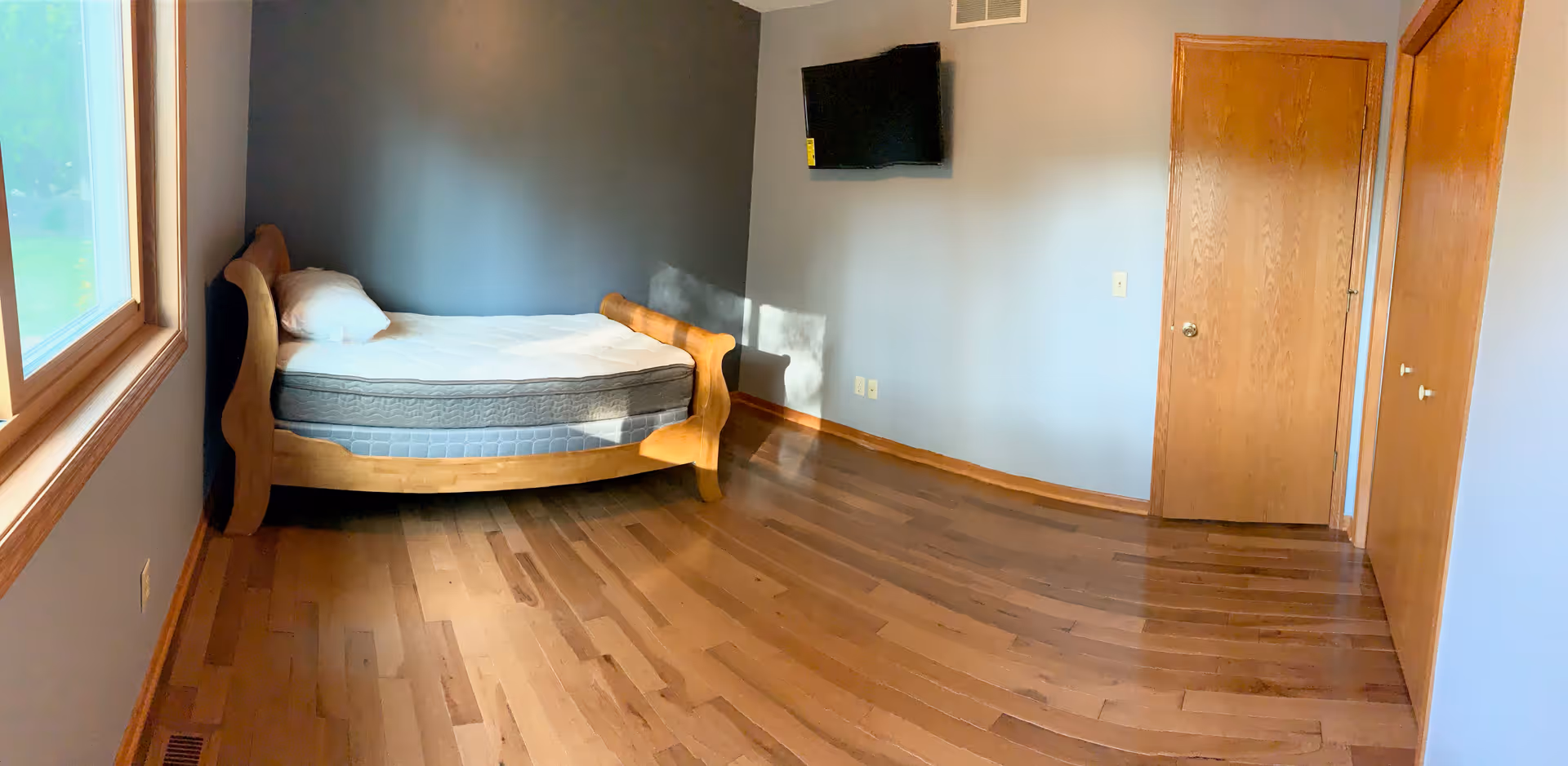 A bedroom with a curved wooden bed, blue walls, a window, mounted TV, and hardwood floor.