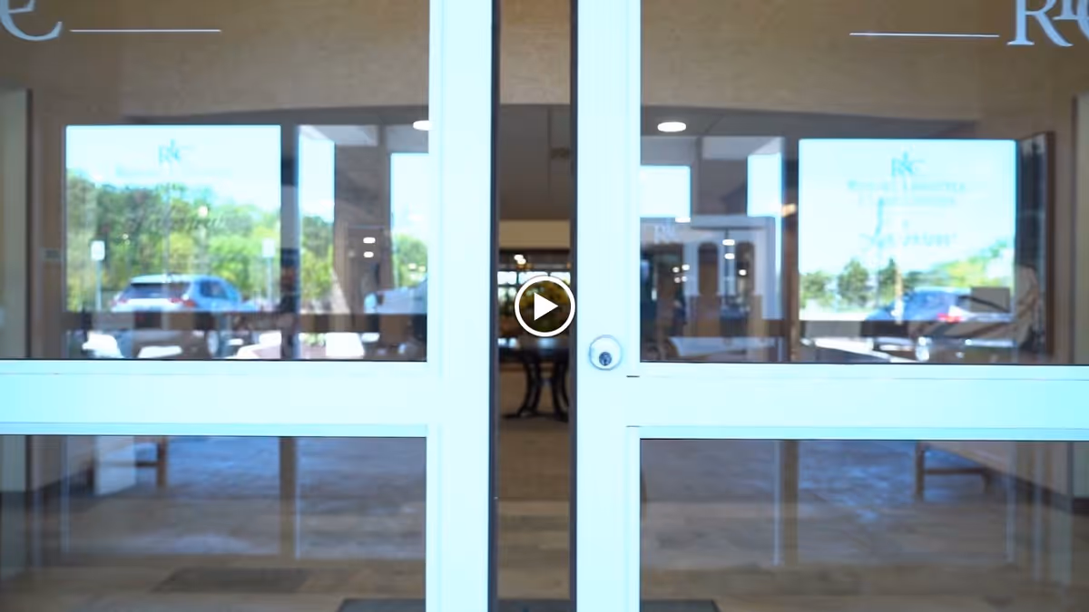 Glass double doors at the entrance of a building with reflections of parked cars and greenery outside. Inside, a hallway with tables and chairs is visible.