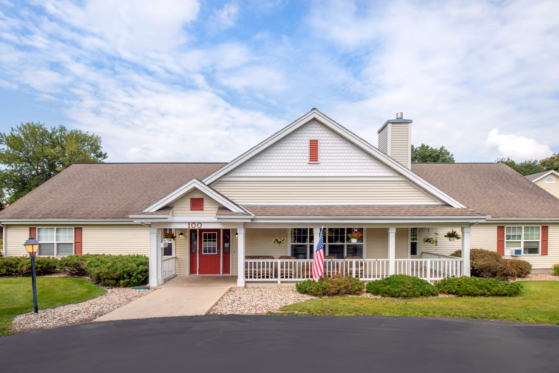 Front exterior view of a single-story building with beige siding and a brown roof. The building has a covered porch with white railings, hanging flower pots, and an American flag. The entrance features double red doors with the number 100 above them. There are bushes and a lamp post in the landscaped front yard under a partly cloudy sky.