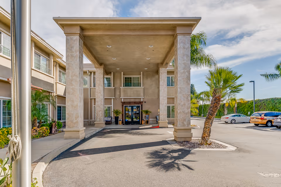 Senior living facility entrance with porte-cochère, tall columns, palm trees, and parked cars.