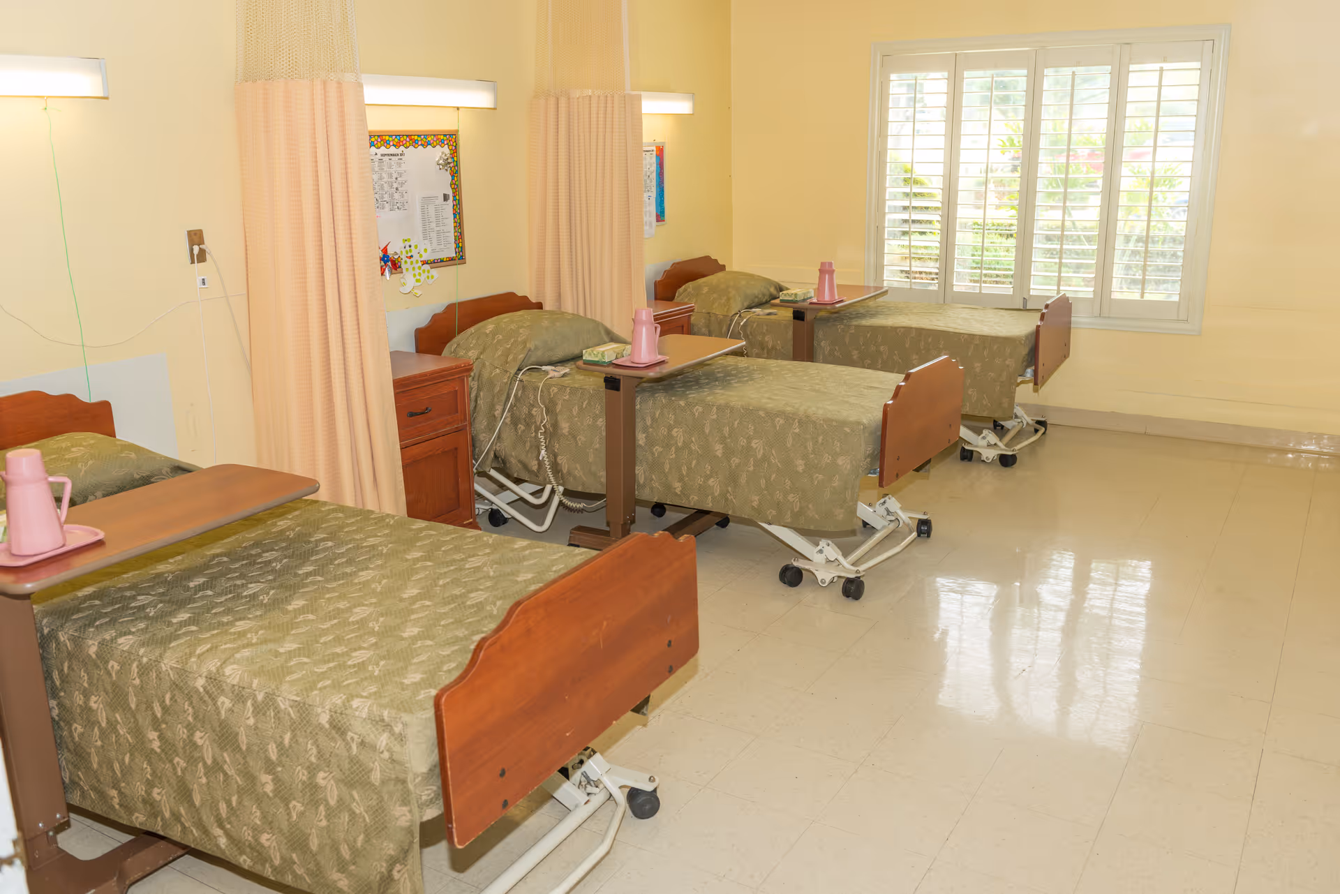 A room with three hospital-style beds arranged in a row, each with green patterned bedspreads and a pink water pitcher on a tray table. The beds have wooden headboards and footboards, and there are privacy curtains between them. A window with white shutters lets in natural light, and the floor is tiled and shiny.