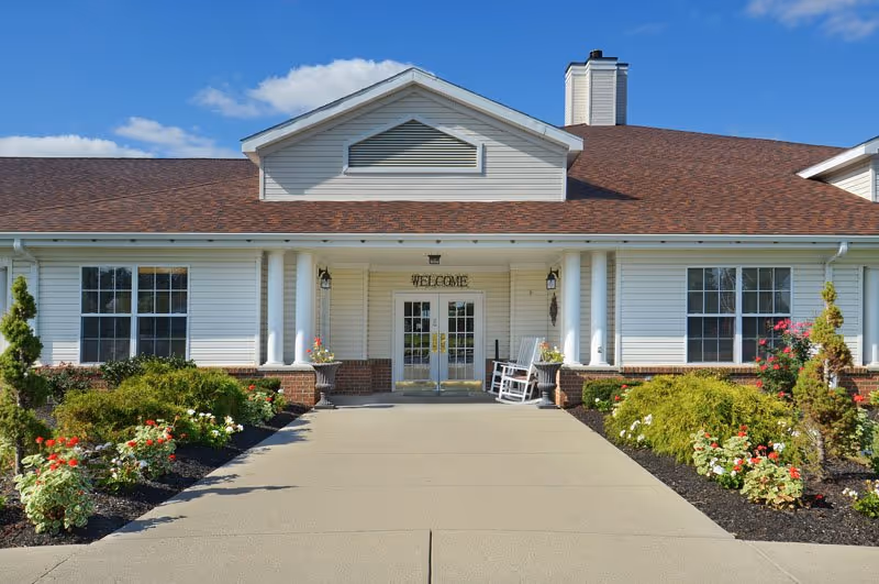 White senior living facility front entrance with seating and manicured landscaping.