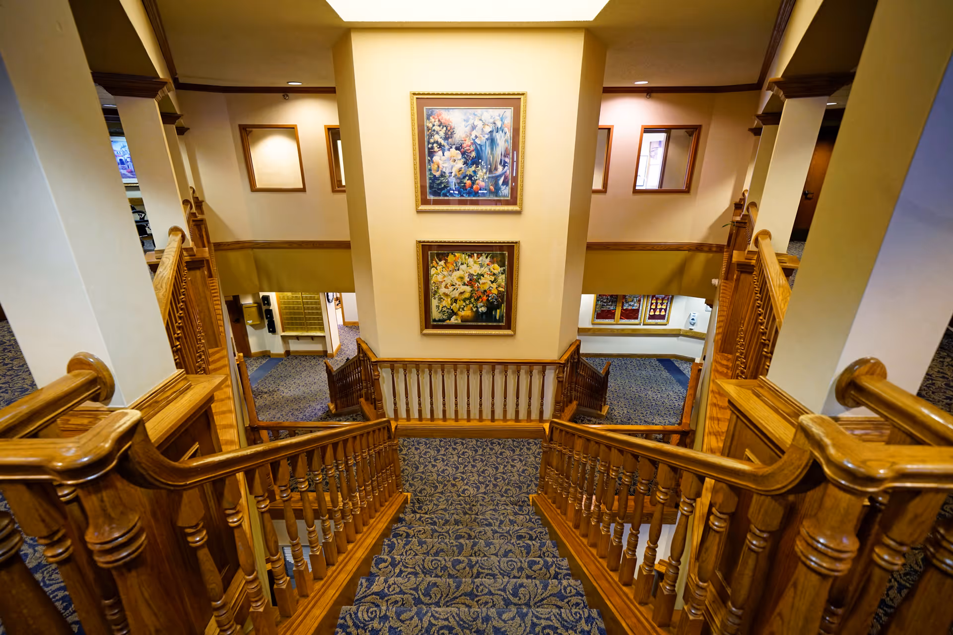 Wooden staircase with blue carpet, railings, and framed paintings in a grand interior.