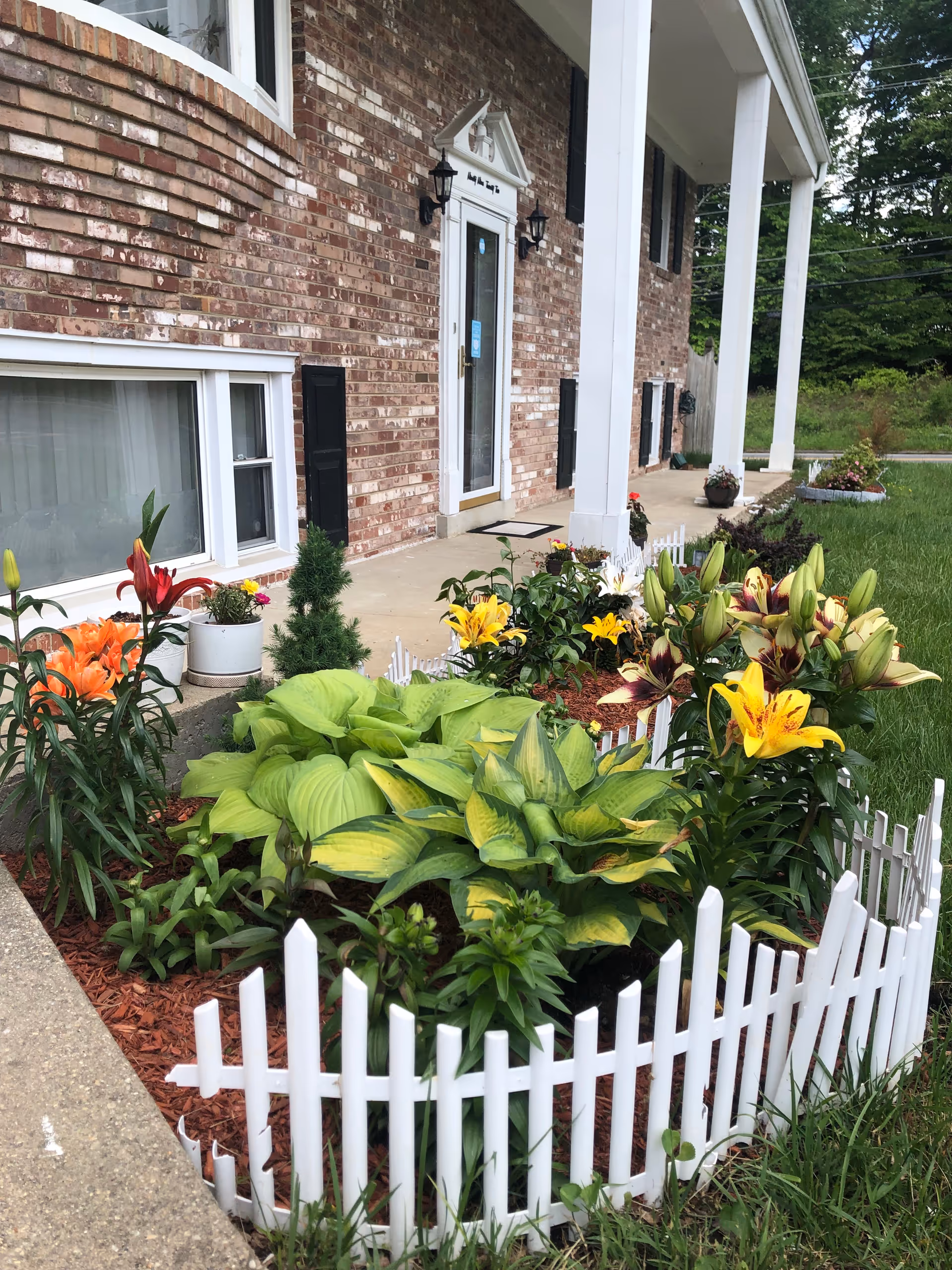 Brick-front building entrance with white columns and a colorful garden bordered by a white picket fence.