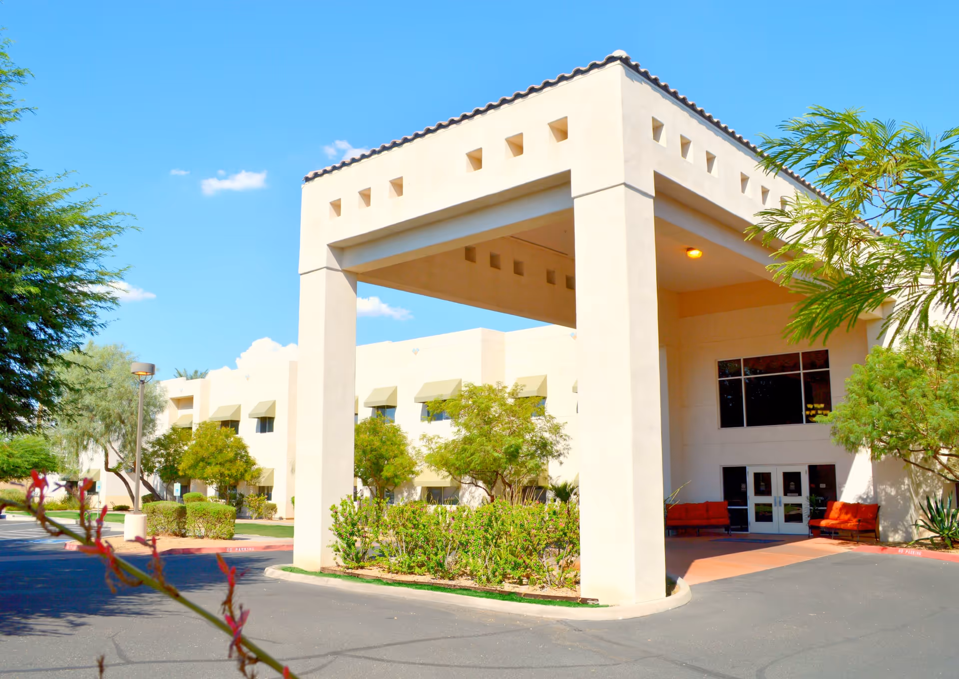 Exterior view of a beige two-story assisted living facility building with a covered entrance supported by large square columns. The building is surrounded by green trees and shrubs under a clear blue sky.