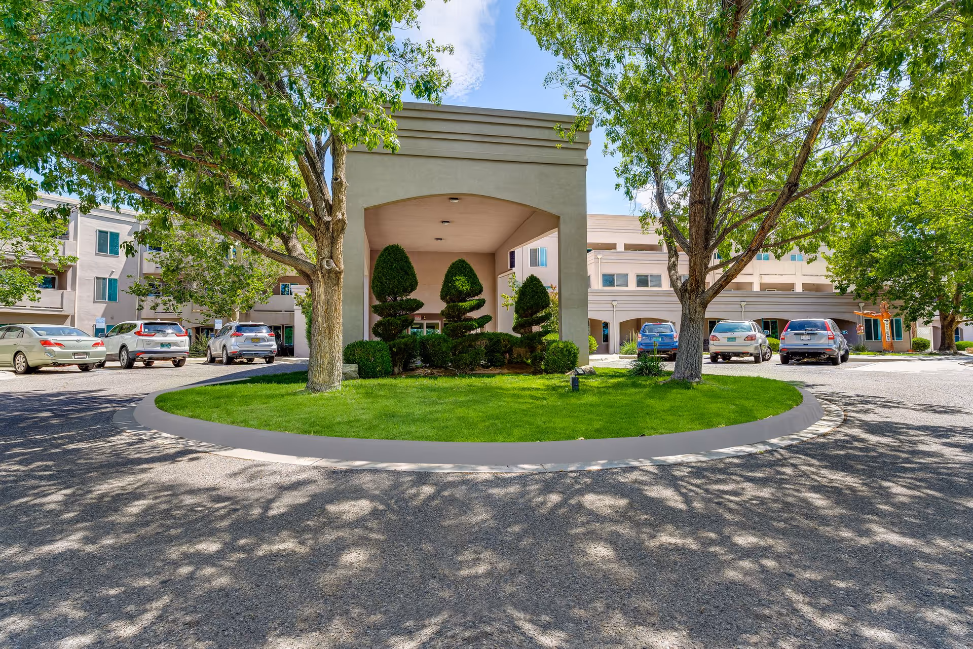 Senior living facility entrance with circular driveway, lush landscaping, and parked cars.