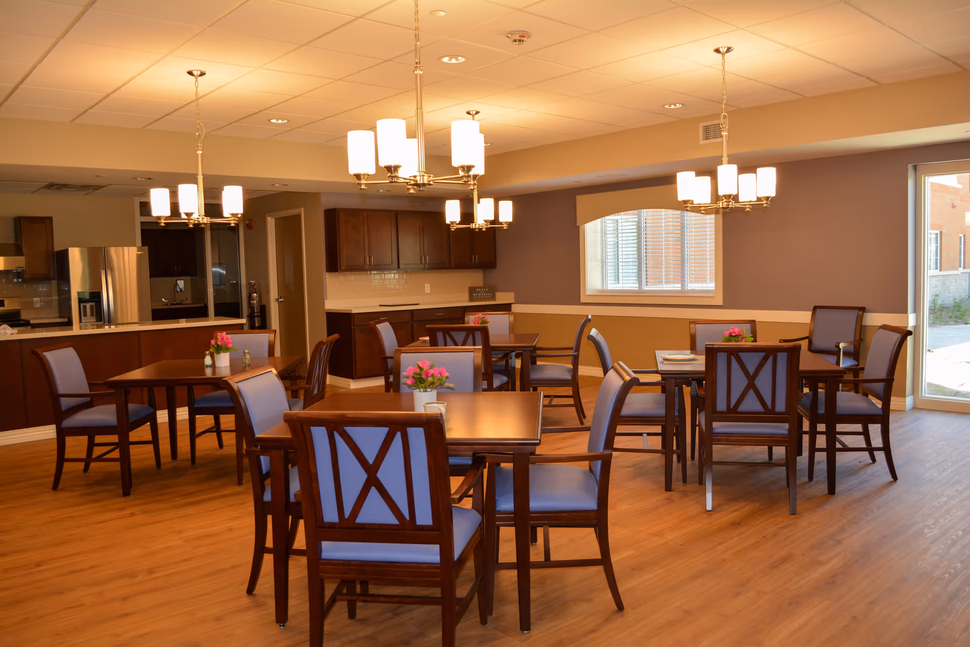 Dining area with wooden tables and blue-upholstered chairs, chandeliers overhead, and a serving counter/kitchen in the background.