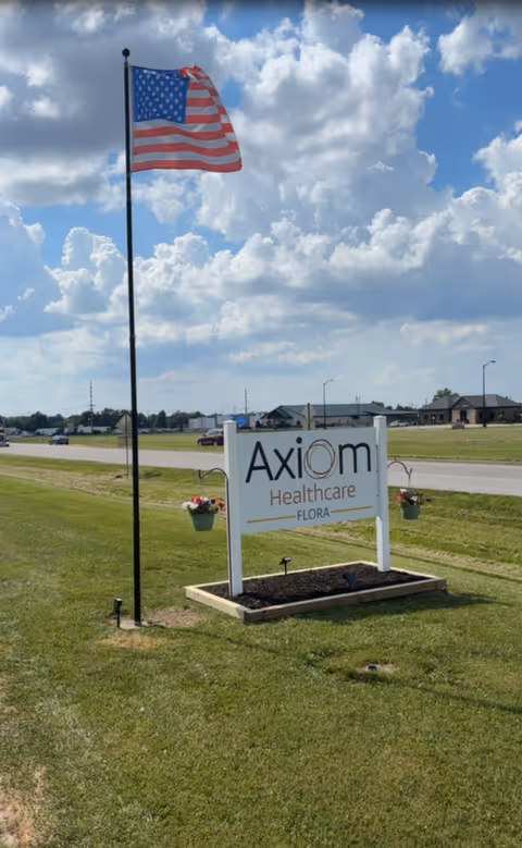 A lawn-mounted sign reading "Axiom Healthcare Flora" next to an American flagpole with buildings and a cloudy sky in the background.