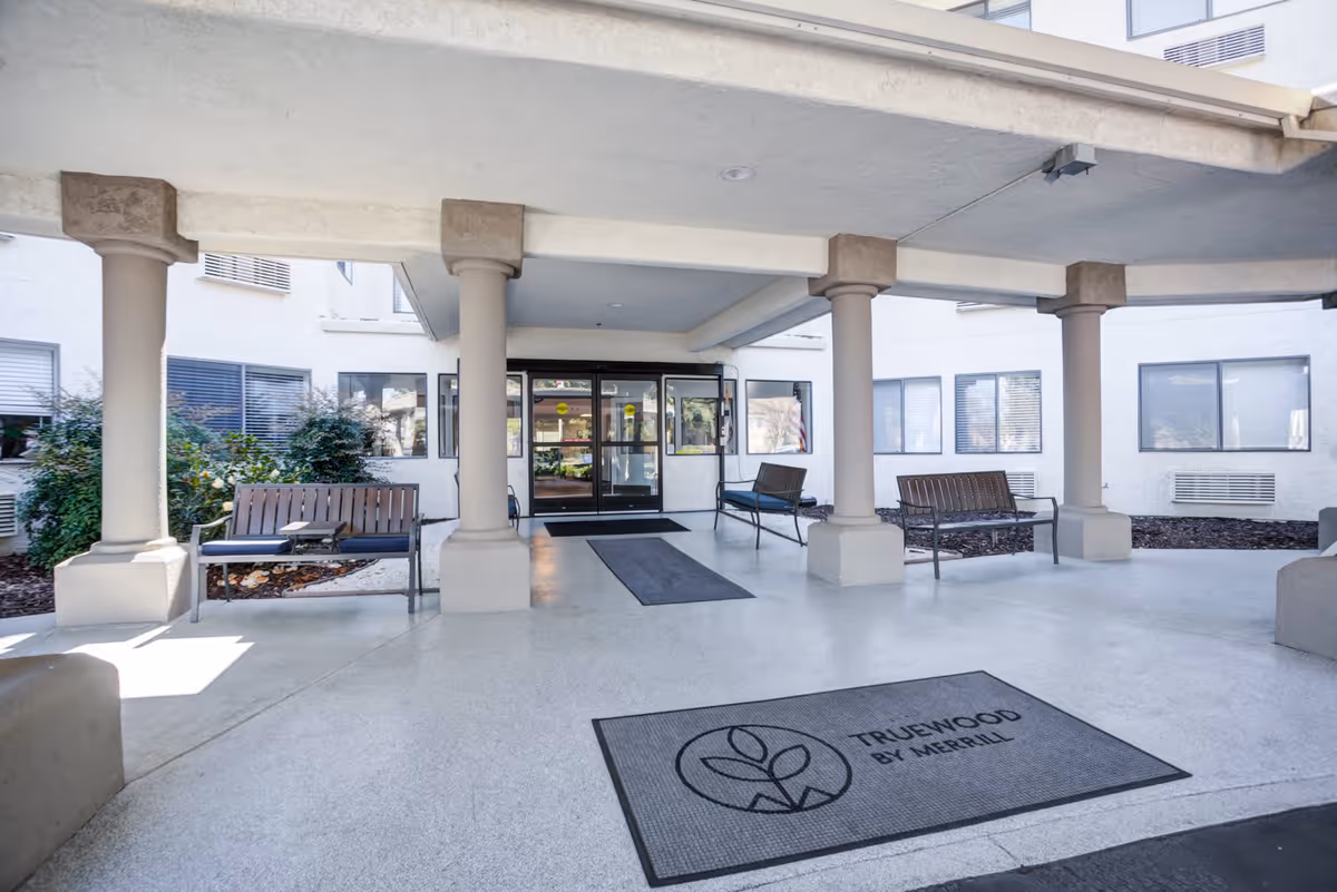Covered entrance area of Truewood by Merrill facility in Modesto with benches on either side and a doormat displaying the facility's logo and name. The entrance has glass doors and is supported by several columns.