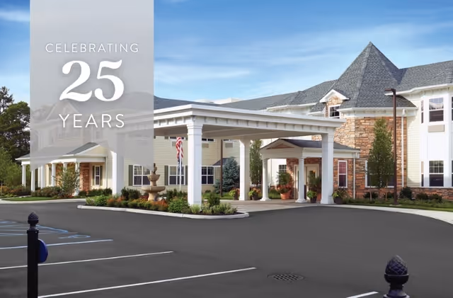 Front entrance of an assisted living building with a covered porte-cochere, landscaped beds and empty parking lot, overlaid by a 'Celebrating 25 Years' banner.
