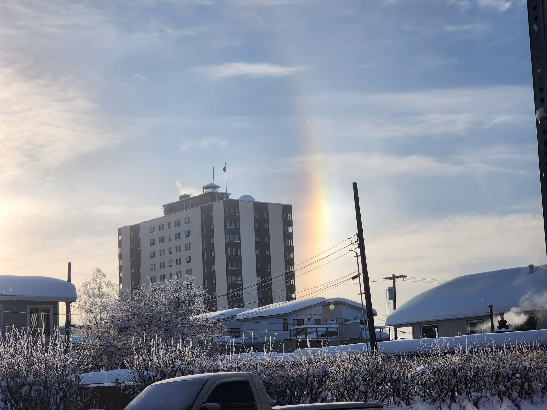 Snowy street scene with a tall building and a rainbow behind.
