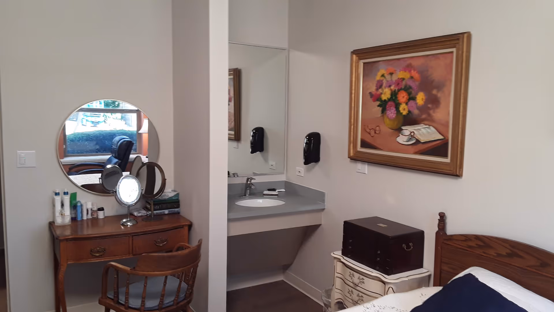 A bedroom with wooden dresser, round mirror, sink vanity, framed painting, and bed.