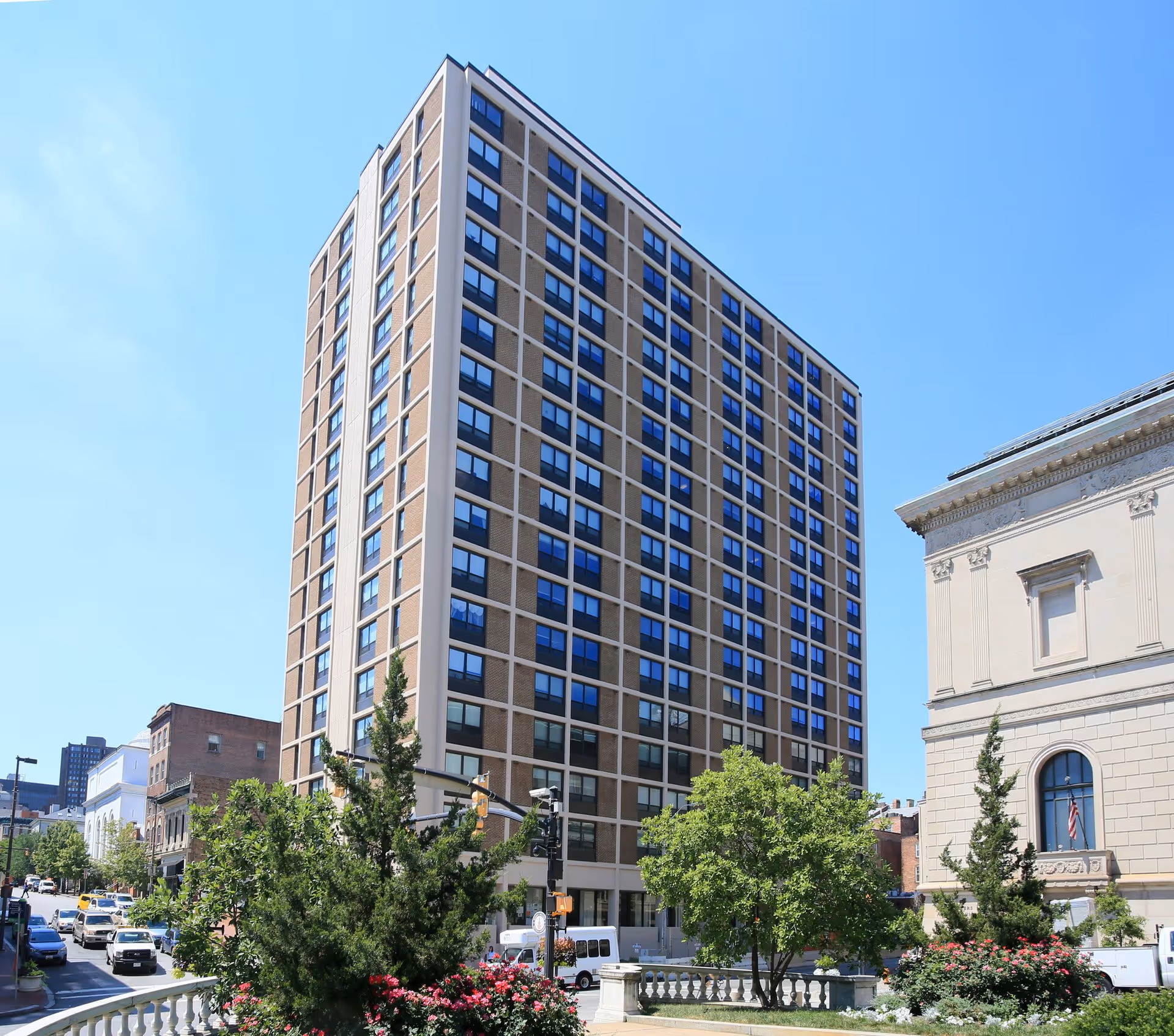 Tall multi-story apartment building with trees and a street in front under a clear blue sky.