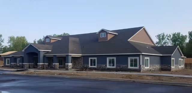 Exterior view of a single-story building with a dark roof, gray walls, and stone accents at the base. The building has multiple windows and a covered entrance. The surrounding area includes a paved road and some trees in the background.