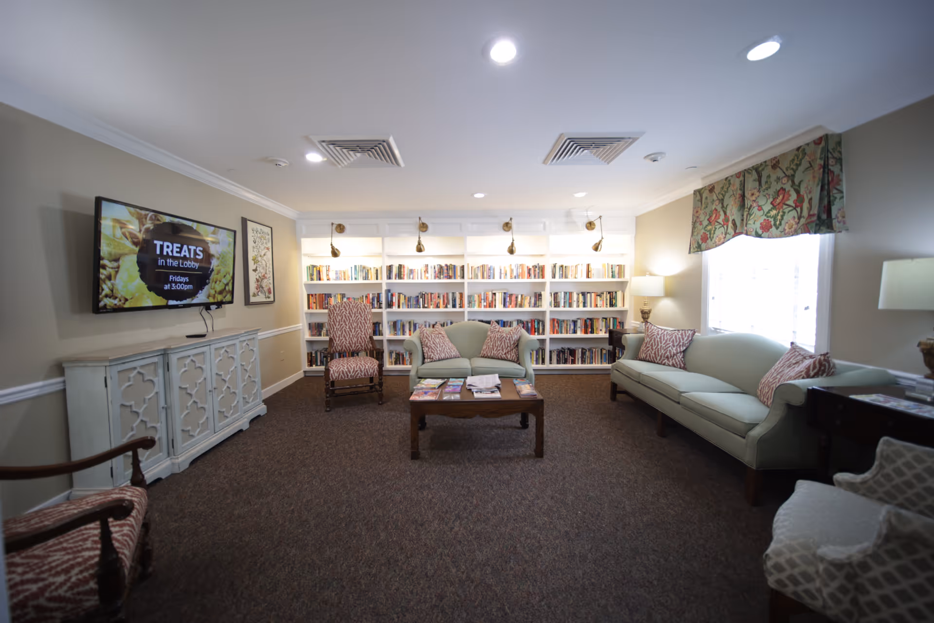 Cozy senior living lounge with sofas, chairs, bookshelves, and a central coffee table.