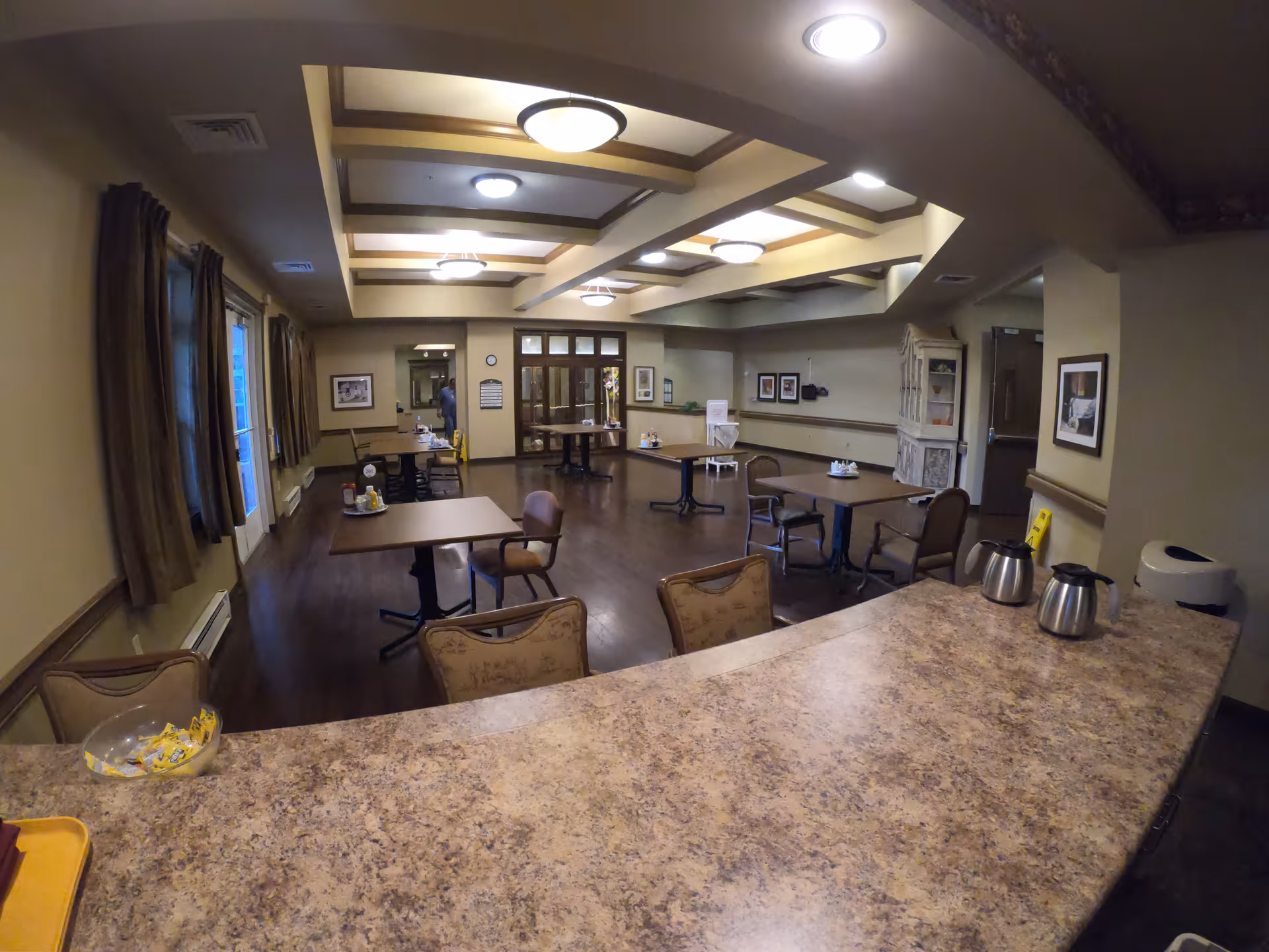 Interior view of a dining room in a senior living facility with several tables and chairs arranged neatly. The room has a coffered ceiling with multiple light fixtures, windows with brown curtains on the left, and framed pictures on the walls. A countertop with coffee pots and a bowl of sugar packets is visible in the foreground.