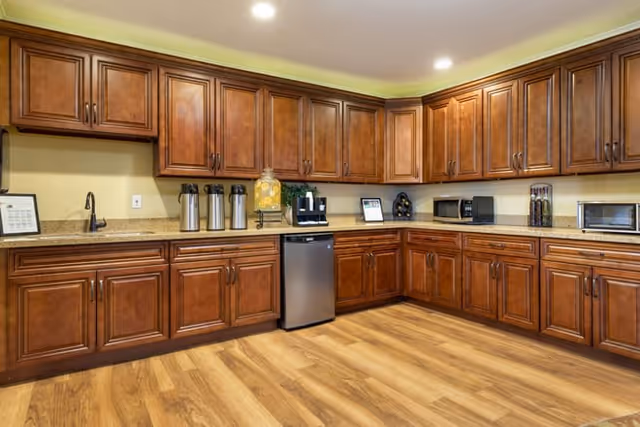 A spacious kitchen area with wooden cabinets and drawers lining the walls. The countertop holds several appliances including a coffee machine, a microwave, a toaster oven, and beverage dispensers. The floor is made of light wood, and the ceiling has recessed lighting.