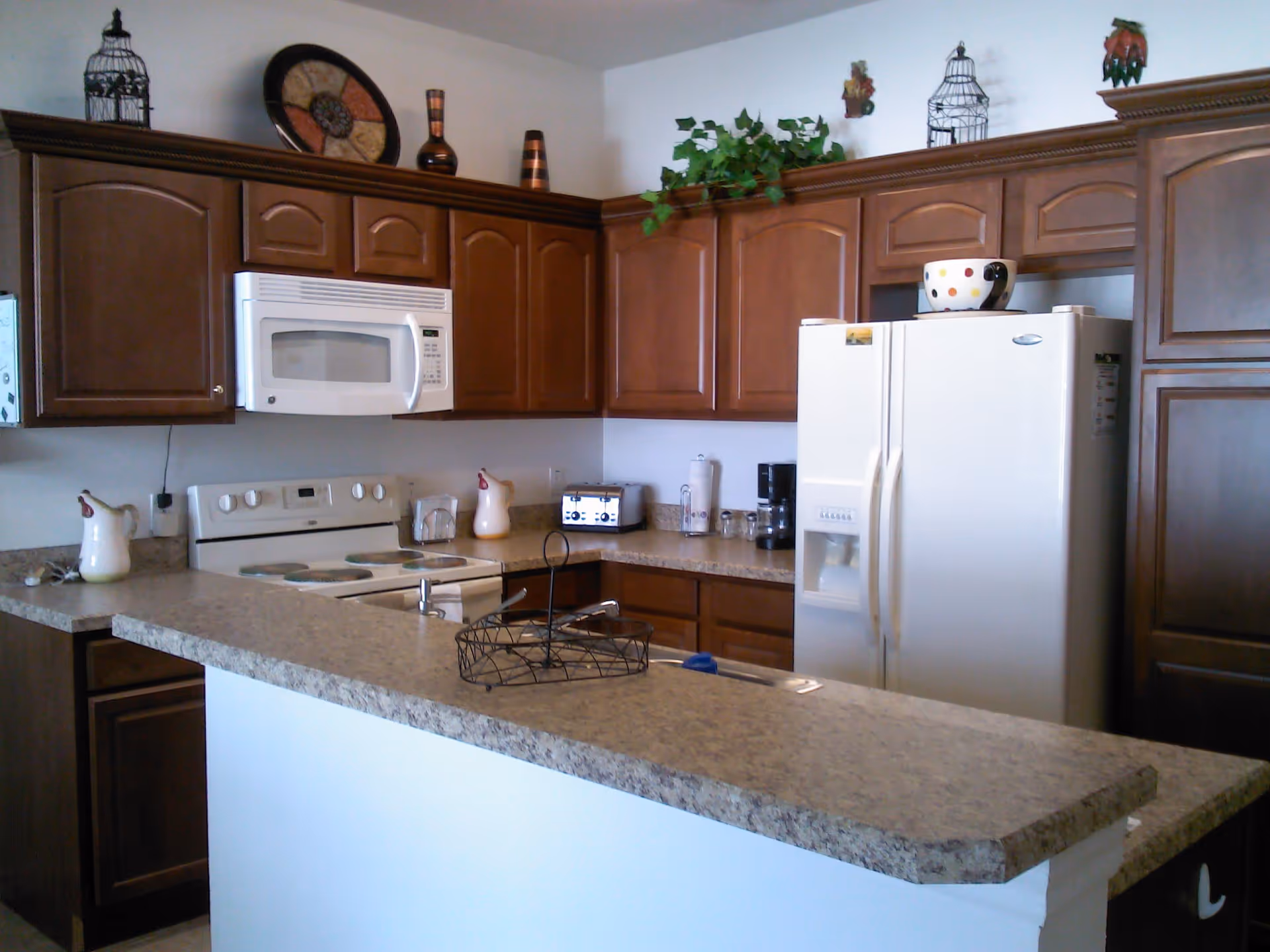 A kitchen with brown wooden cabinets, a white microwave above a white stove, a white refrigerator, and a countertop with a wire basket. Decorative items including vases, a plate, and plants are placed on top of the cabinets.