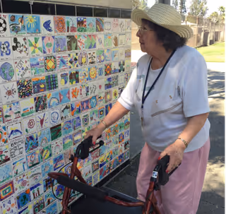 An elderly woman wearing a sun hat examines a colorful tiled wall outdoors.