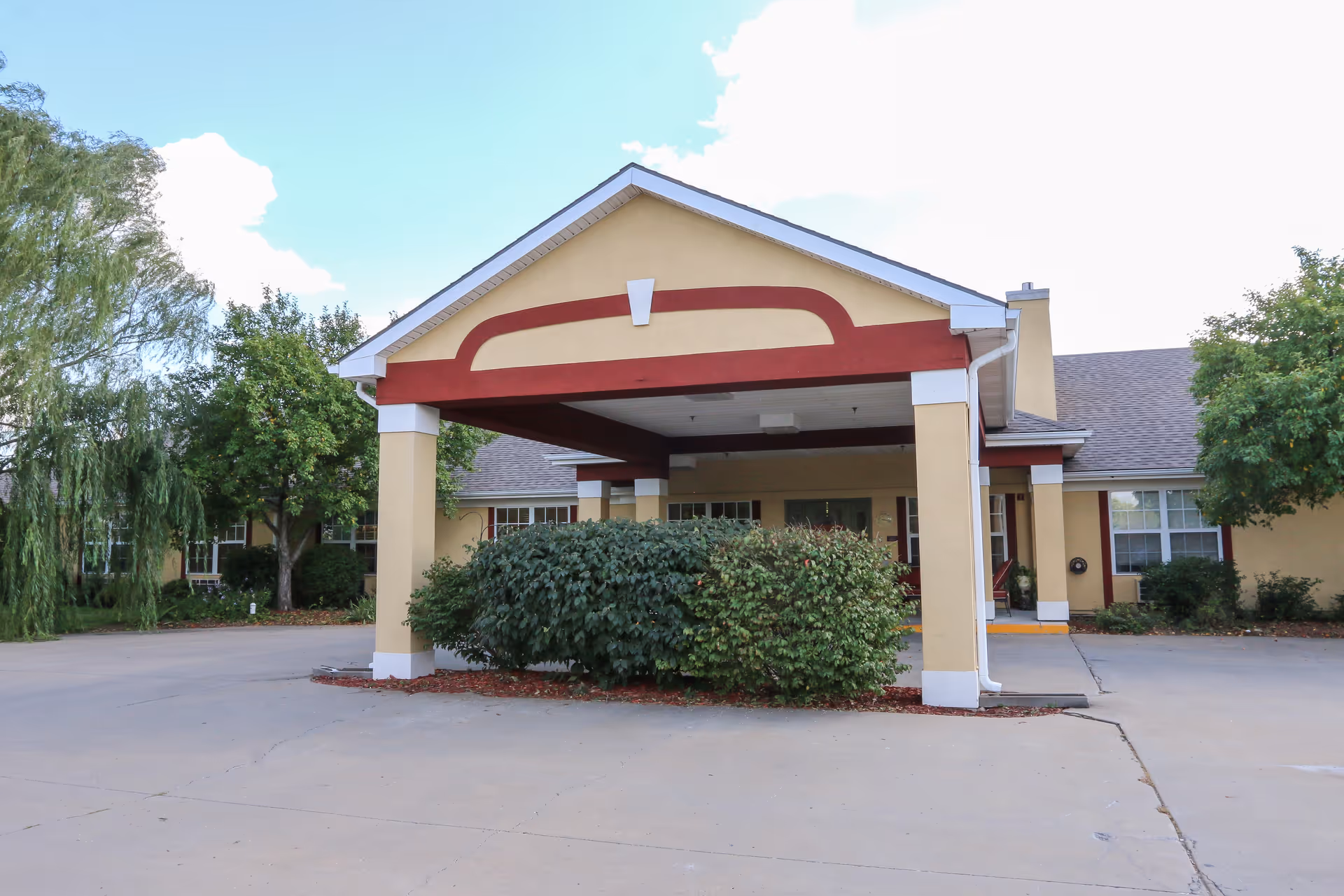 Covered porte-cochere entrance of a single-story building with shrubs and a driveway.