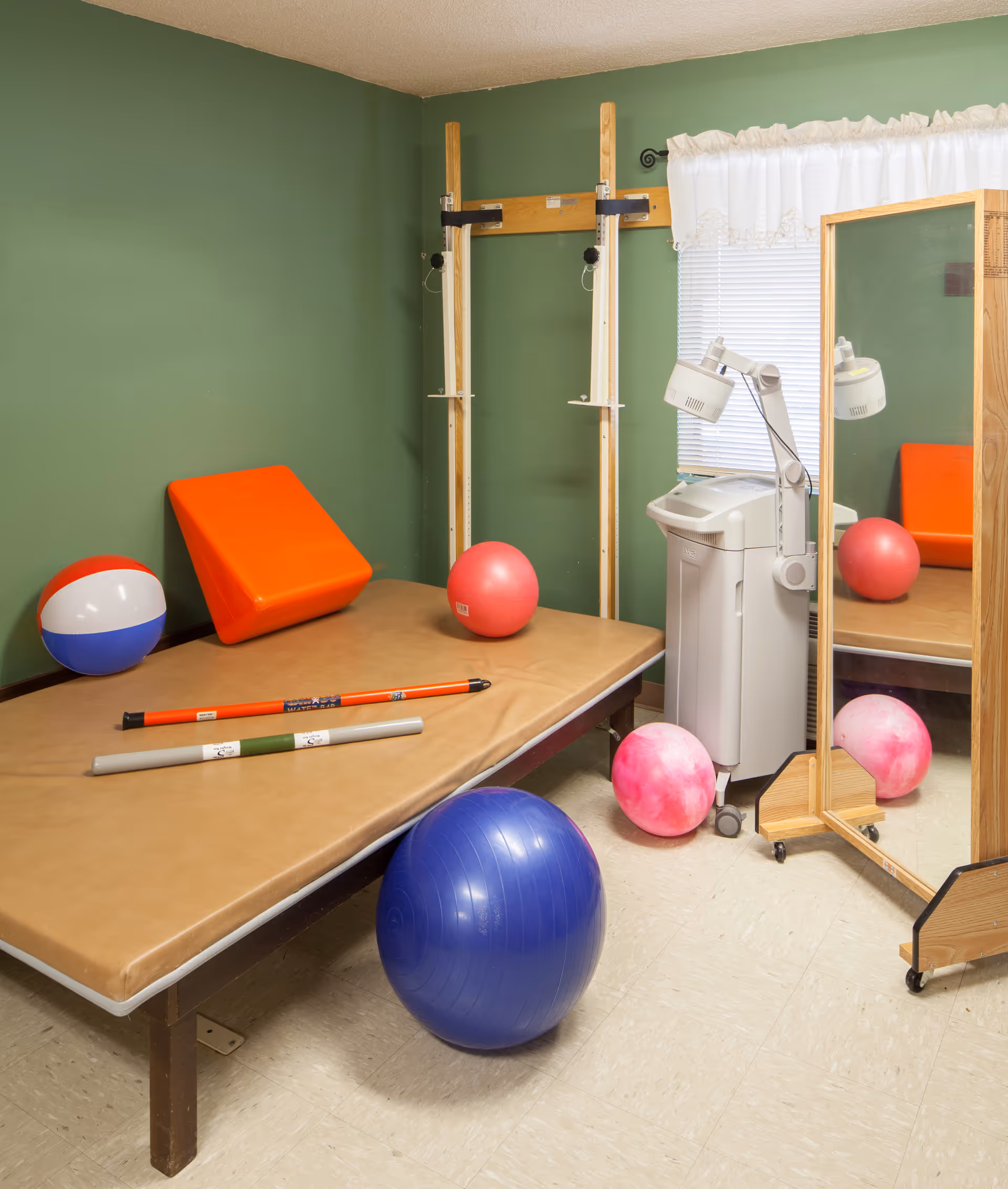 Green-walled therapy room with a padded treatment table, exercise balls, and rehabilitation equipment.