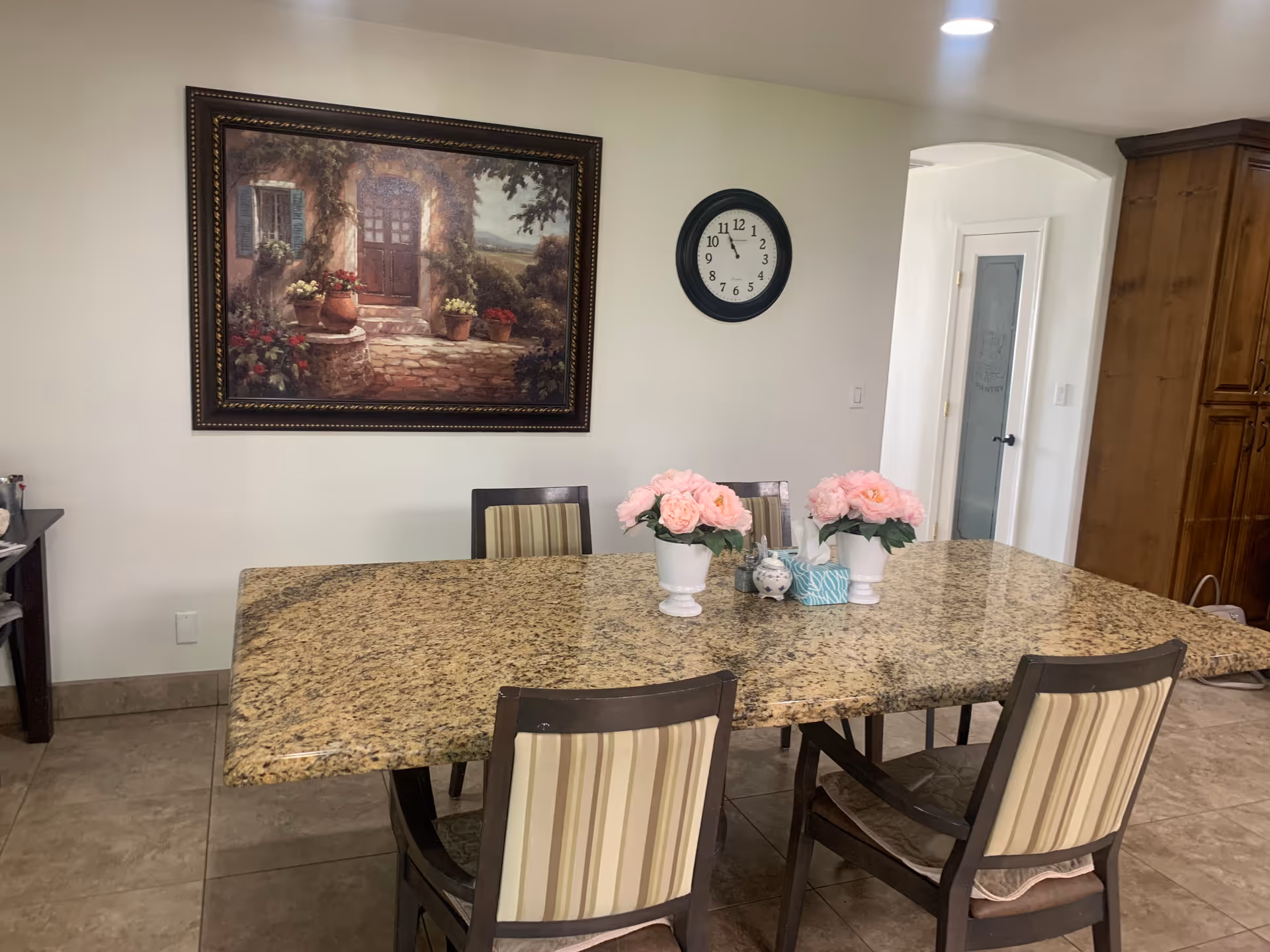 Dining area with a large granite table and four chairs, pink floral centerpieces, a wall clock and framed painting.
