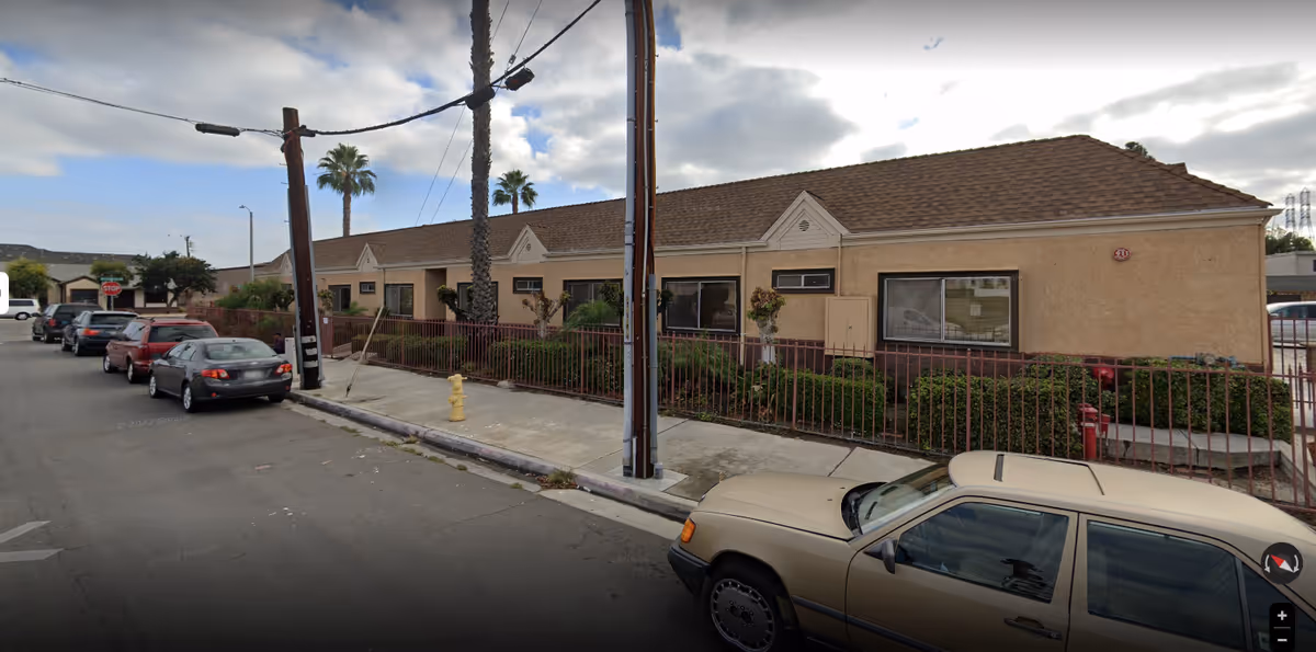 Single-story stucco building with a fenced front yard, palm trees, and cars parked along the street.