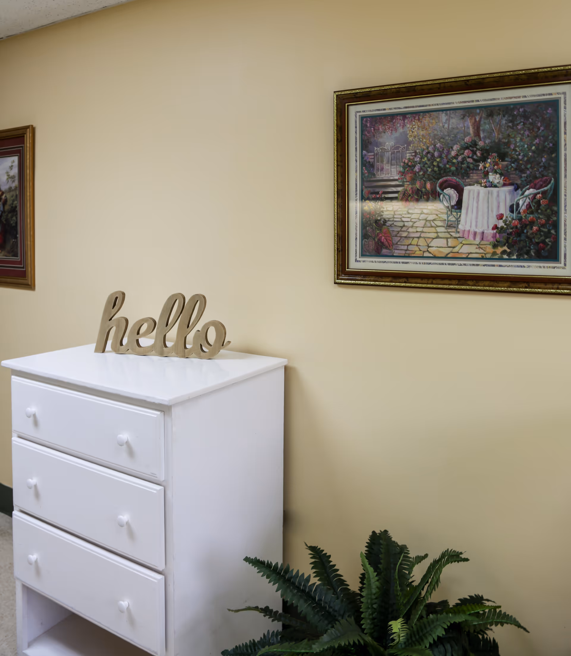 A white three-drawer dresser topped with a decorative "hello" sign, framed artwork on a beige wall, and a potted fern on the floor.