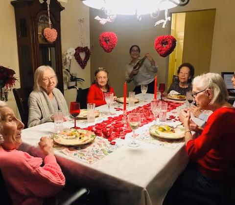 Several elderly women seated around a dining table decorated with rose petals, red candles and heart decorations, with a staff member standing in the background.