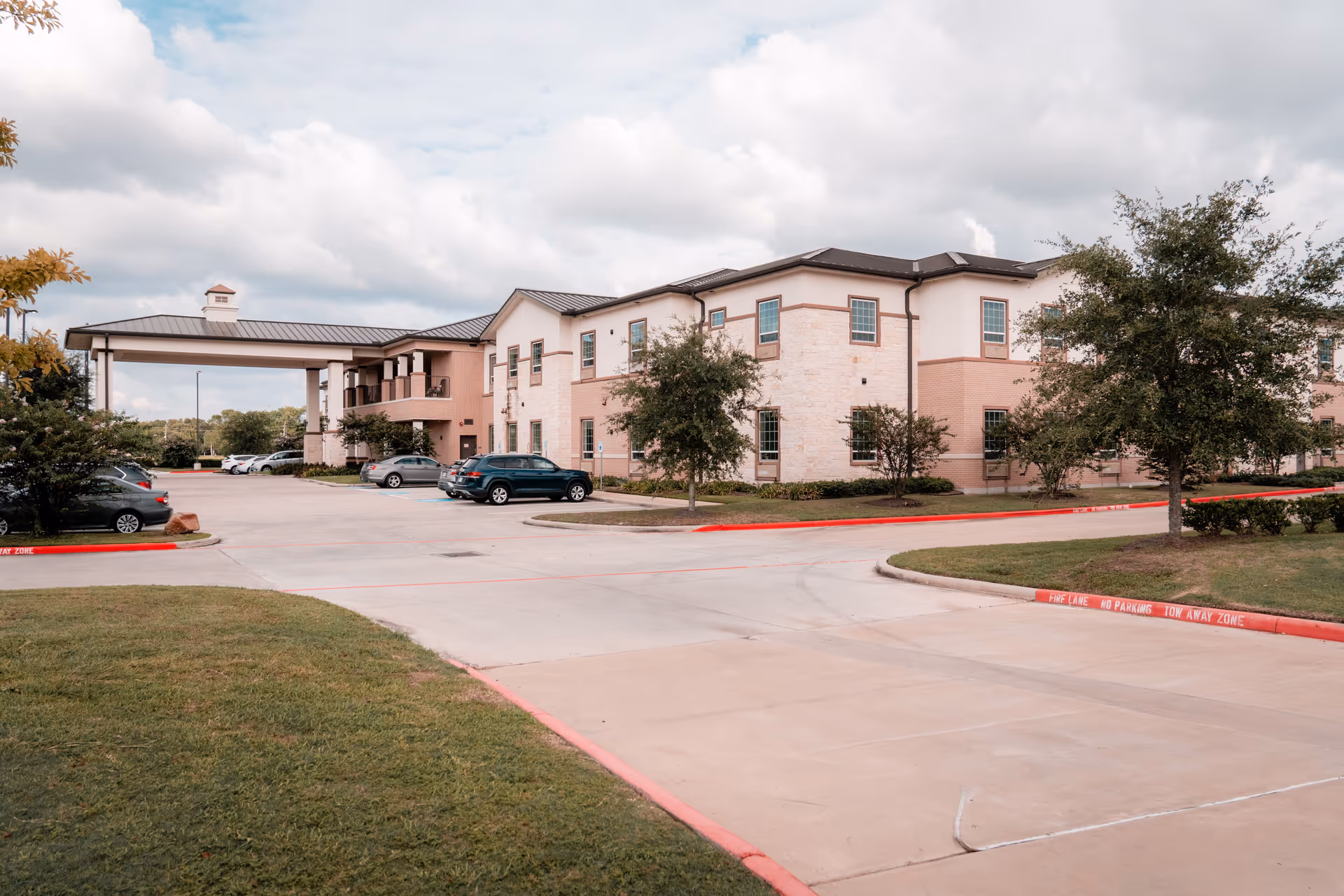 Two-story senior living building with a covered entrance, parking area, and landscaped grounds under a cloudy sky.