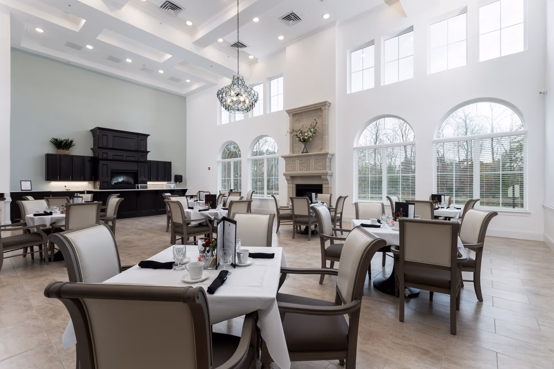 Bright, airy dining room with multiple set tables, high ceilings, large arched windows, chandelier and a fireplace.