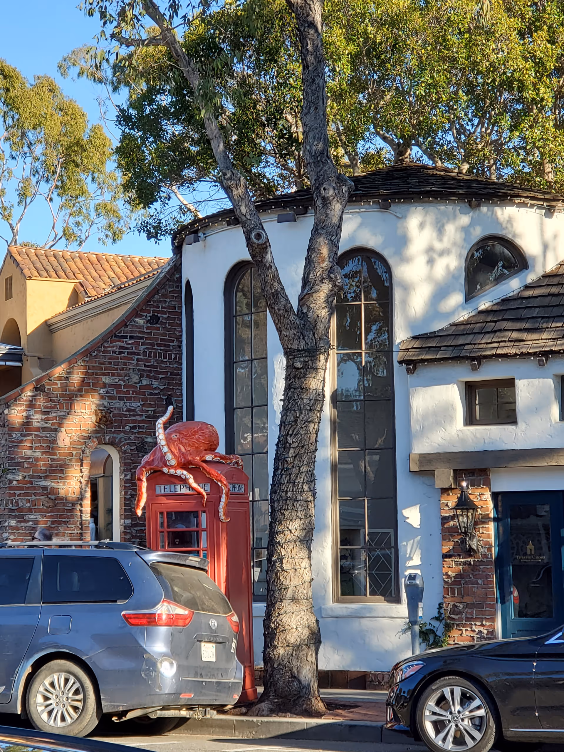 Whitewashed building facade with tall arched windows, a red telephone booth, a tree, and parked cars.