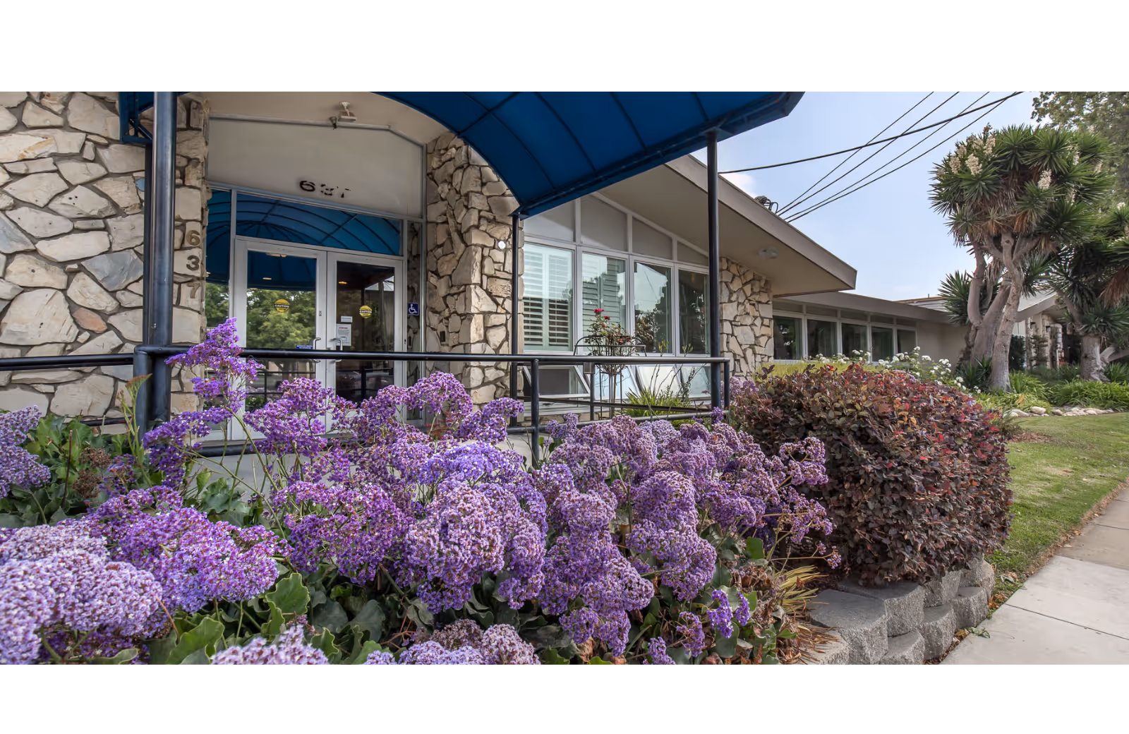 Stone-front entrance of Windsor Gardens Rehabilitation Center with blue awning and purple flowers in bloom